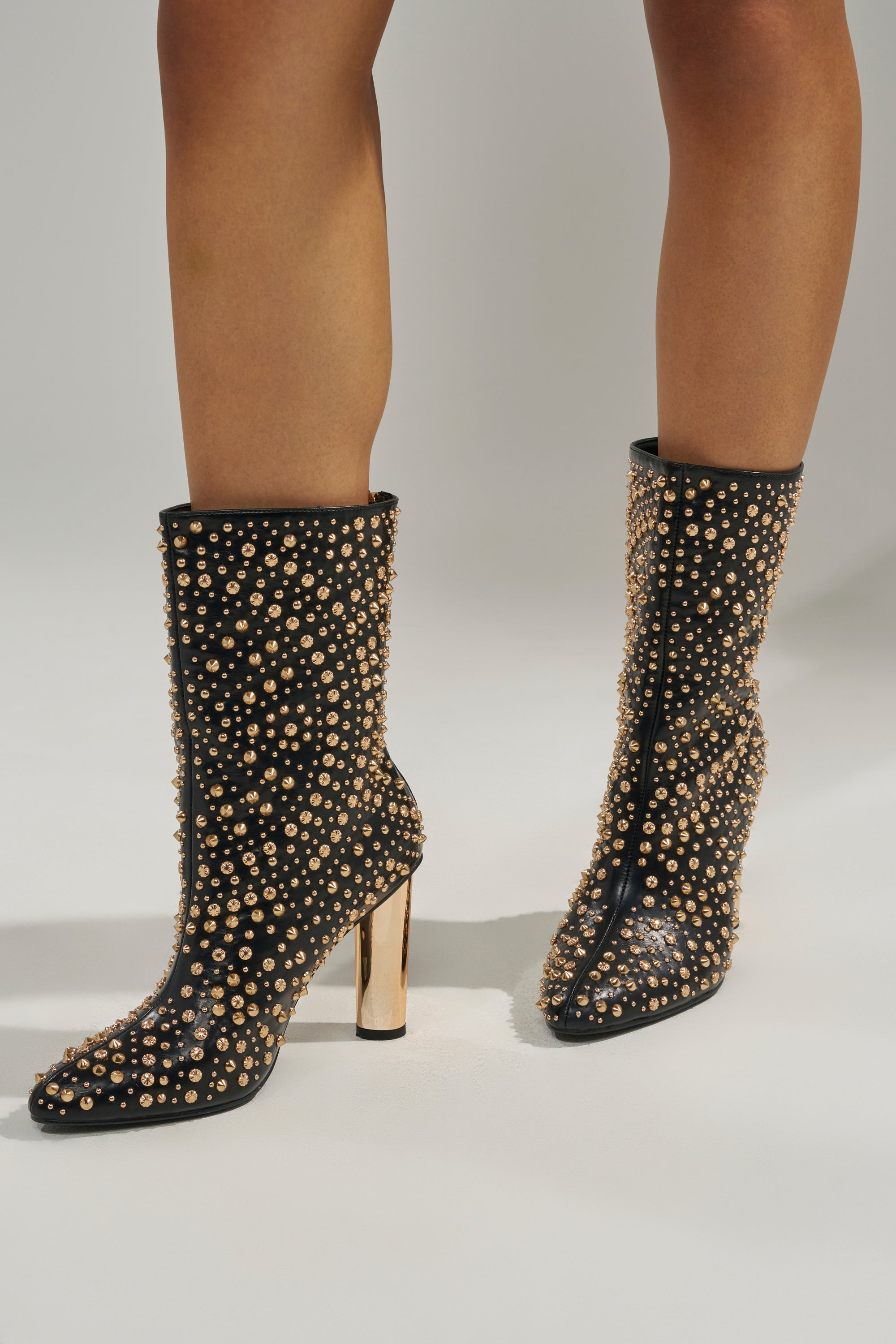 The AZALEA WANG GREHGAN BLACK EMBELLISHED CHUNKY HEEL BOOTIE features black uppers, gold stud embellishments, pointed toes, and metallic gold block heels, seen worn by a person standing on a light surface.
