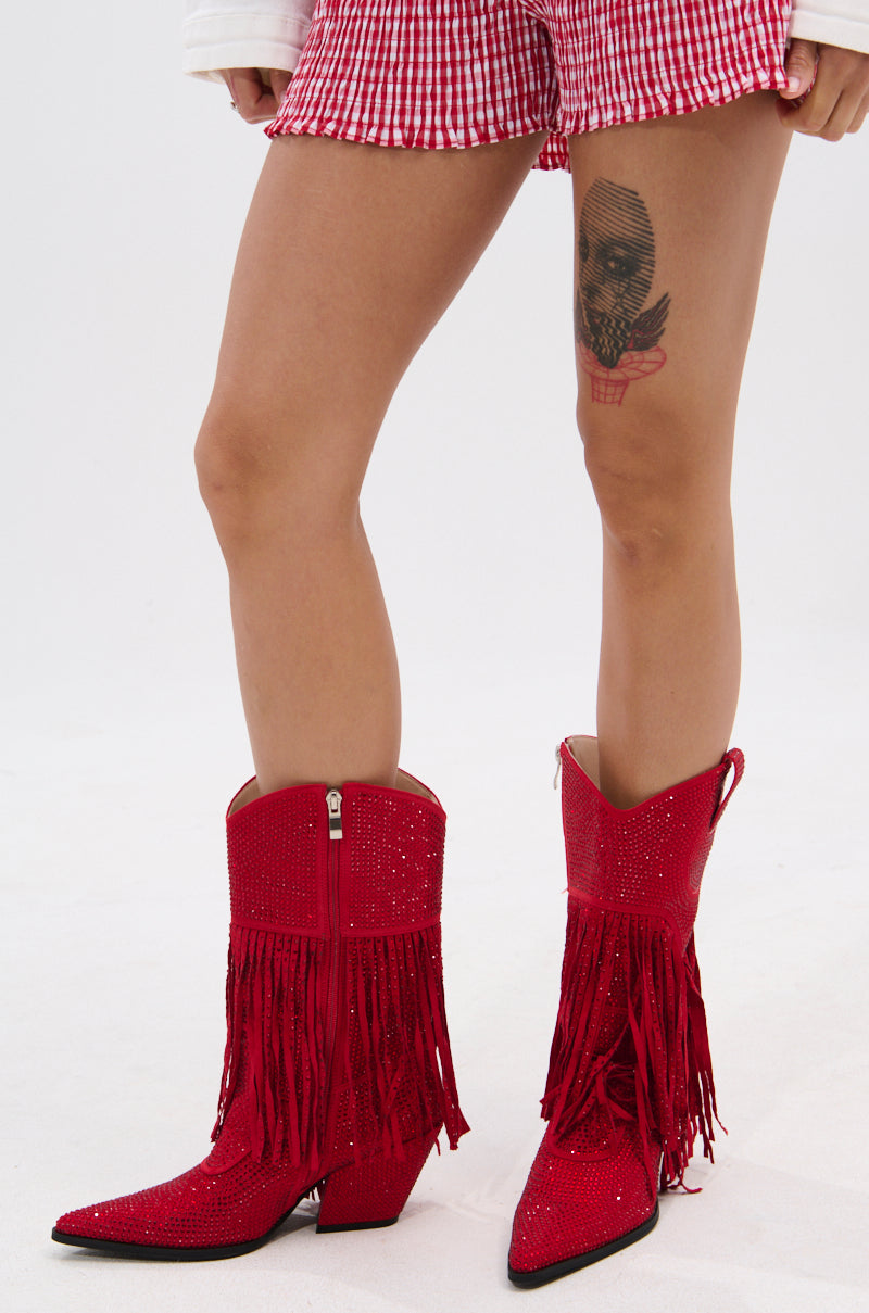 Wearing red, checkered shorts and AZALEA WANG GREIA RED RHINESTONE COWBOY BOOTIE with faux suede details, a person with a skull-in-heart thigh tattoo stands against a plain white background.