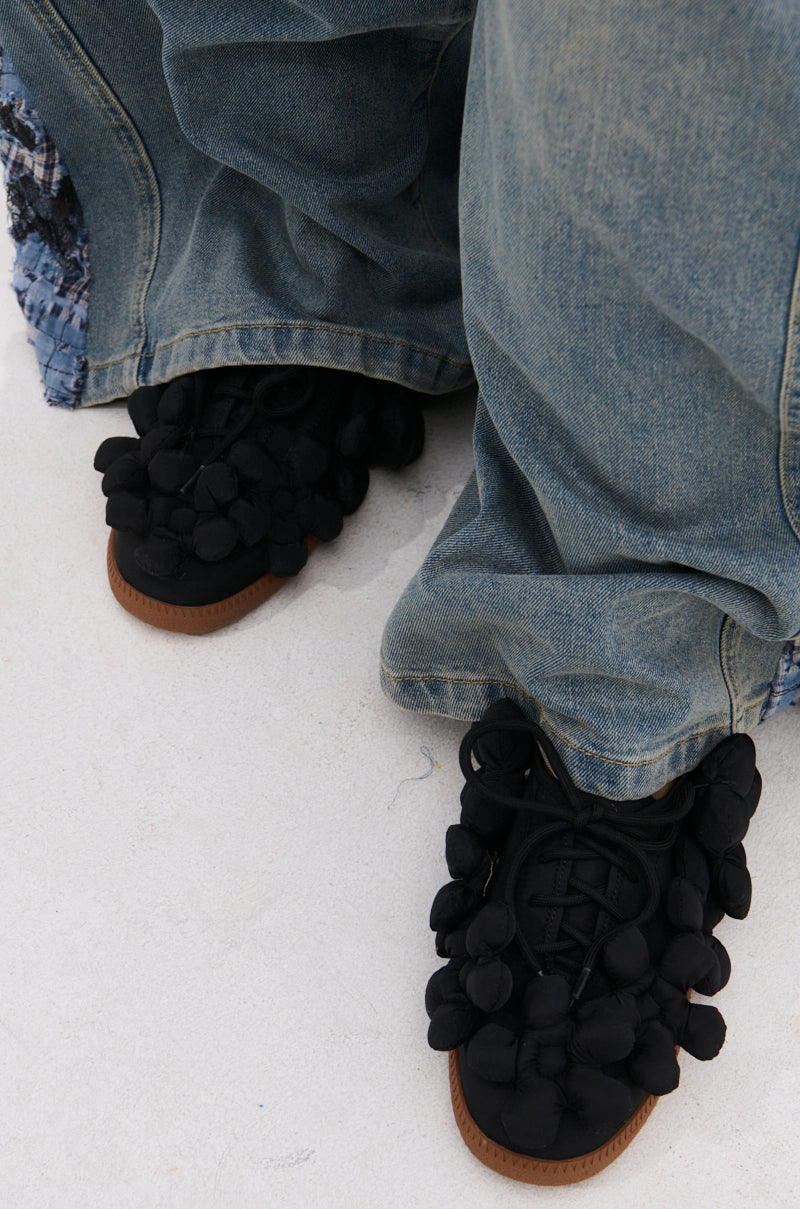 A close-up of someone in wide-legged, faded blue jeans wearing AZALEA WANG GREYI BLACK PUFF FLORAL FLAT SNEAKERS adorned with fabric pom-poms, standing on a light-colored surface.