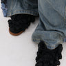 A close-up of someone in wide-legged, faded blue jeans wearing AZALEA WANG GREYI BLACK PUFF FLORAL FLAT SNEAKERS adorned with fabric pom-poms, standing on a light-colored surface.