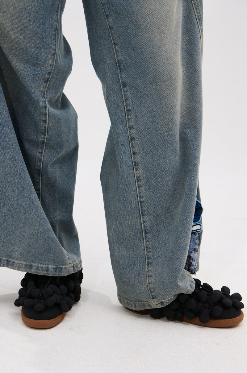 A person wearing loose-fitting blue jeans and AZALEA WANG GREYI BLACK PUFF FLORAL FLAT SNEAKERS stands on a light-colored floor.
