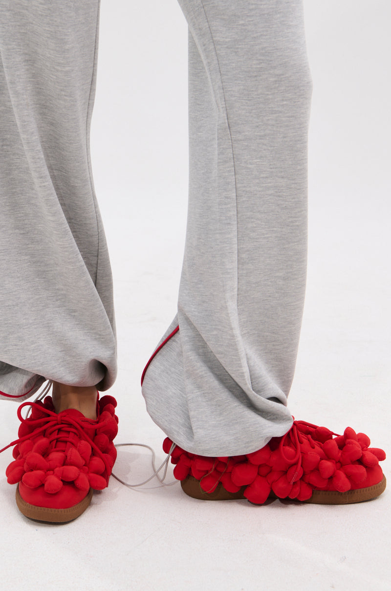 A person in light gray sweatpants wears bold red AZALEA WANG GREYI RED PUFF FLORAL FLAT SNEAKERS with large pom-poms and a textile upper, standing on a light-colored surface.