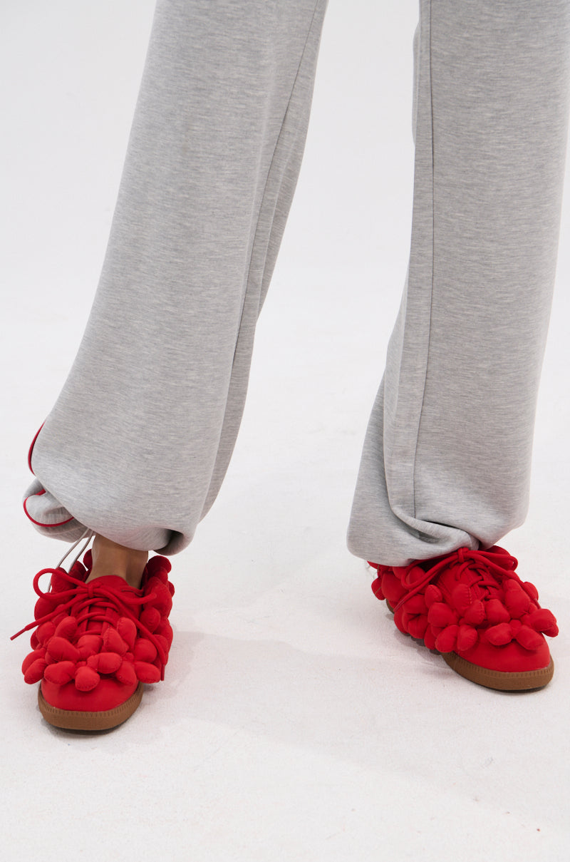 A person in light gray sweatpants wears AZALEA WANG GREYI RED PUFF FLORAL FLAT SNEAKERS with a textile upper and clustered pom-poms, standing on a light-colored floor. Only the lower legs and feet are visible.