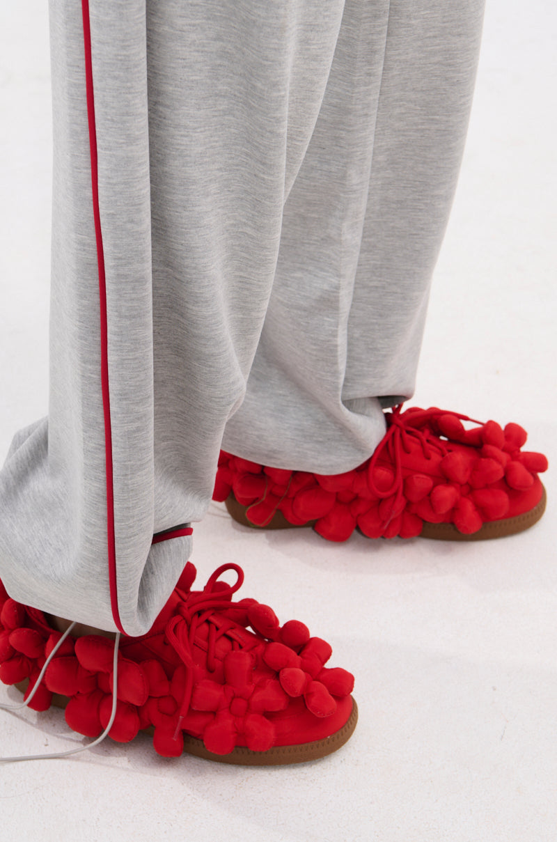 A person in grey pants with a red stripe stands on a light surface, wearing bright red AZALEA WANG GREYI RED PUFF FLORAL FLAT SNEAKERS adorned with playful pom-pom clusters.