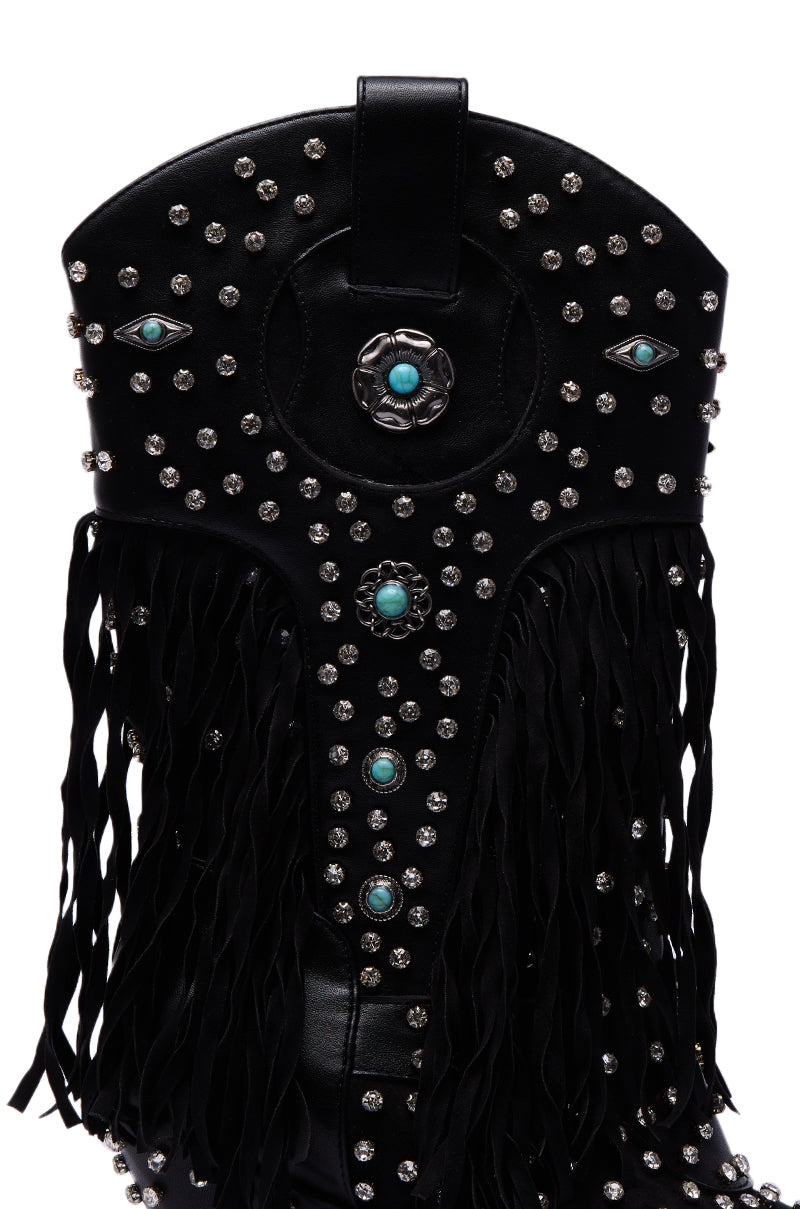 A close-up of the AZALEA WANG GREYSON BLACK BOOTIE in black faux suede, adorned with silver studs, turquoise stones, black fringes, two eye-shaped motifs, and a central circular floral accent.