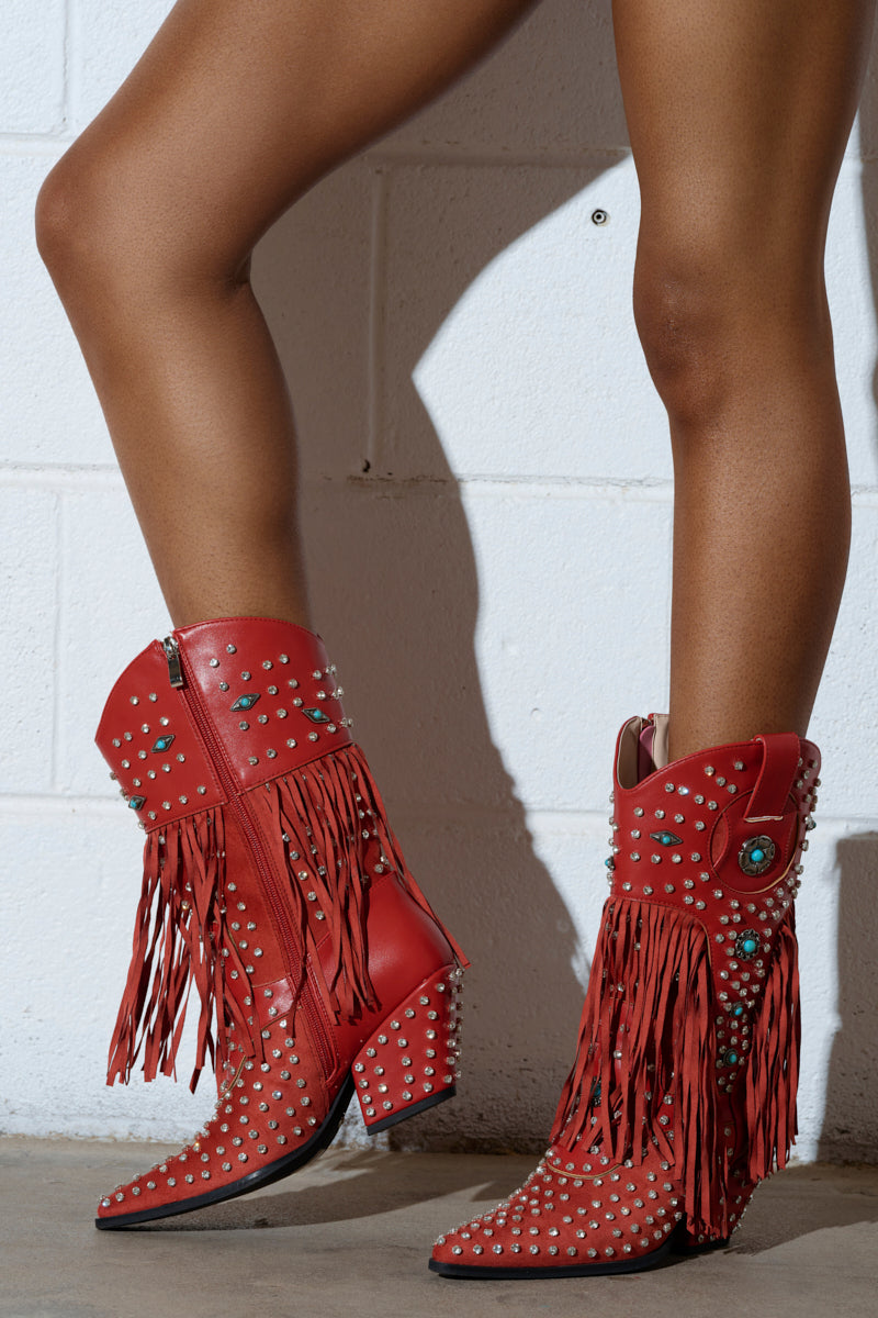 A person wearing AZALEA WANG GREYSON RED BOOT stands against a white brick wall. These Western-inspired boots are adorned with silver studs, turquoise decorative elements, and feature a side zipper paired with a block heel.