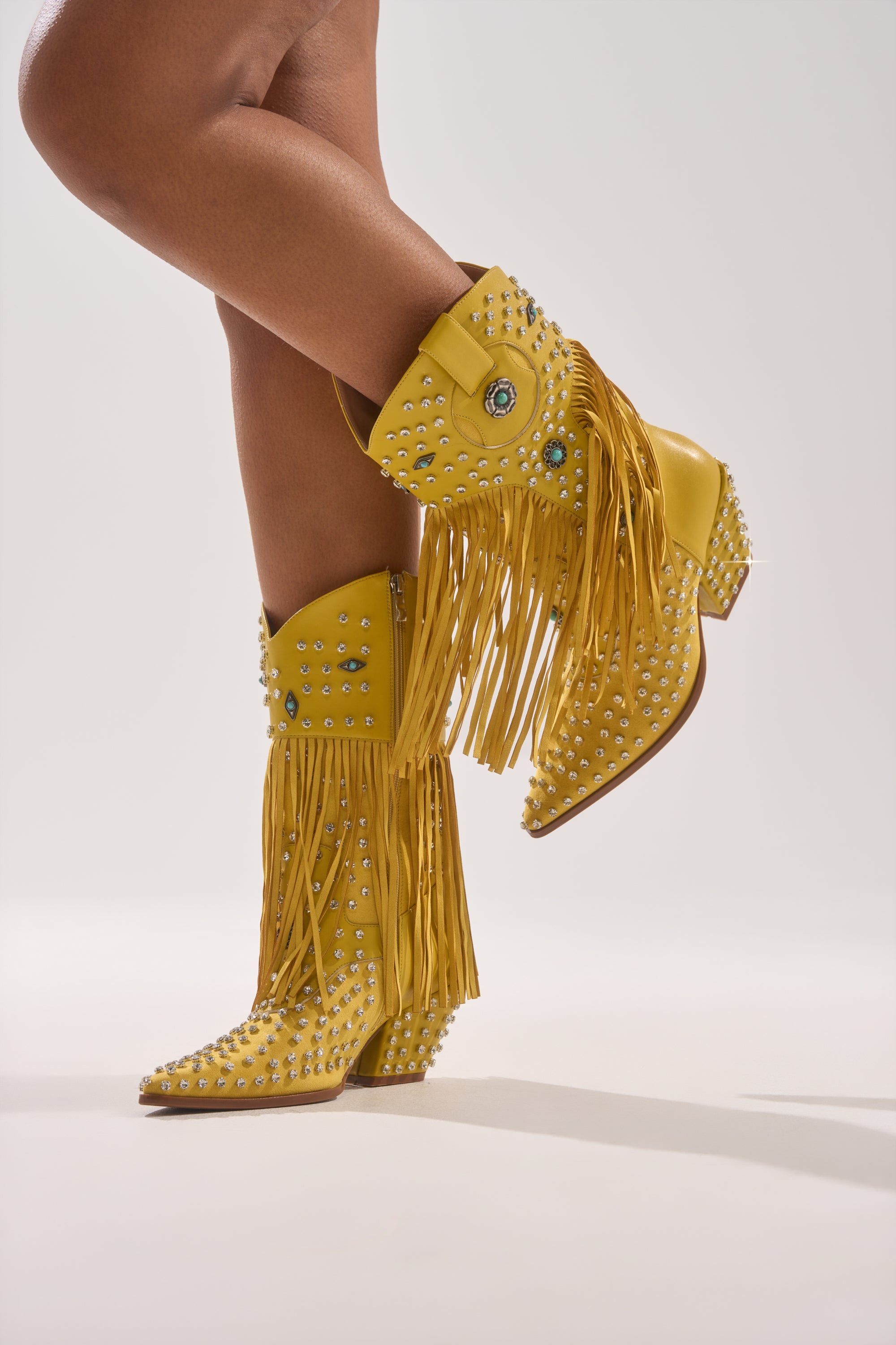 A person wears the AZALEA WANG GREYSON Yellow Rhinestone Embellished Western Bootie, one leg crossed over the other, set against a plain light background.