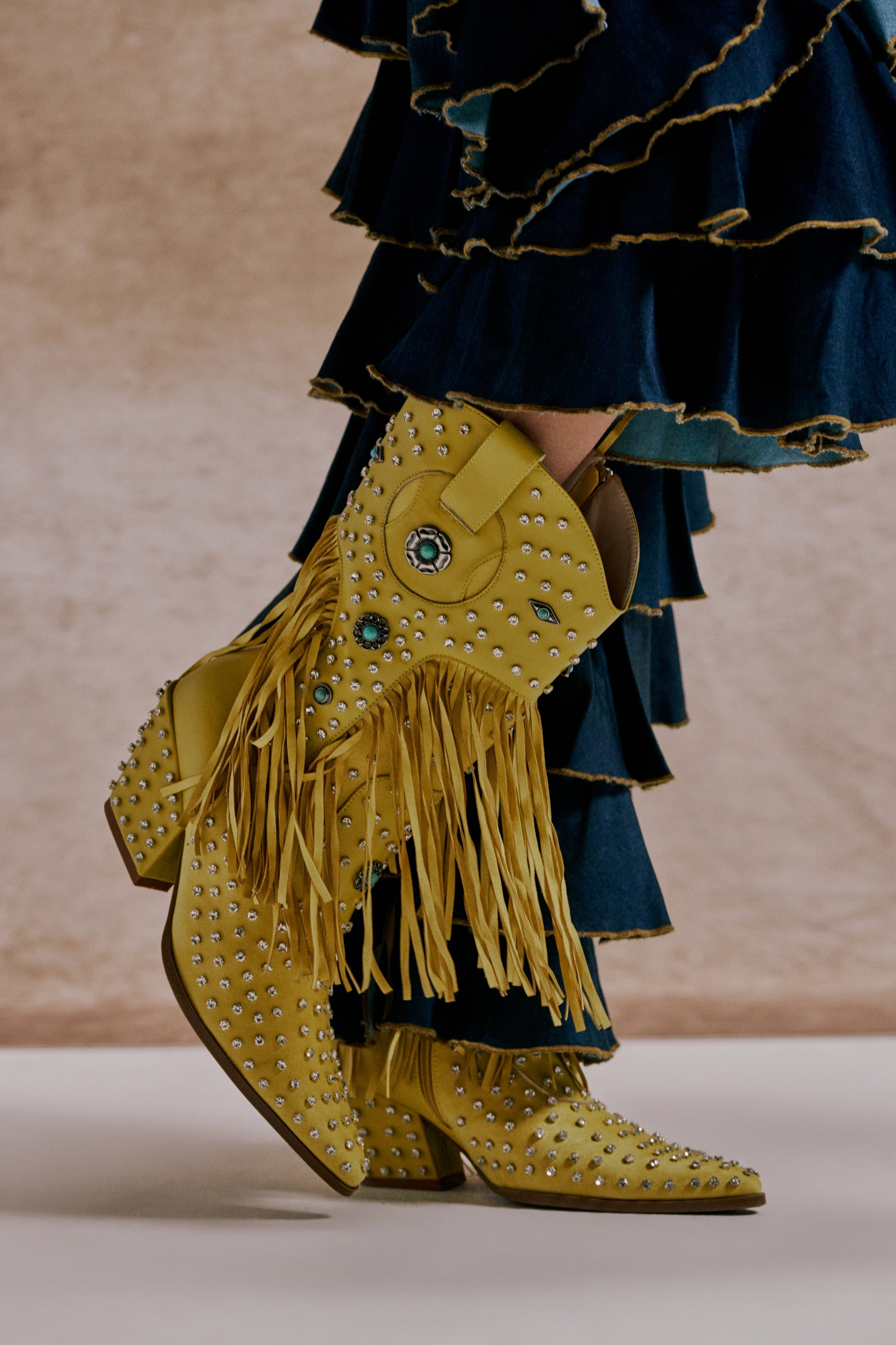 A close-up of the AZALEA WANG GREYSON YELLOW RHINESTONE EMBELLISHED WESTERN BOOTIE, featuring silver studs, turquoise gems, and long fringes, styled with dark blue ruffled pants against a neutral beige background. 0