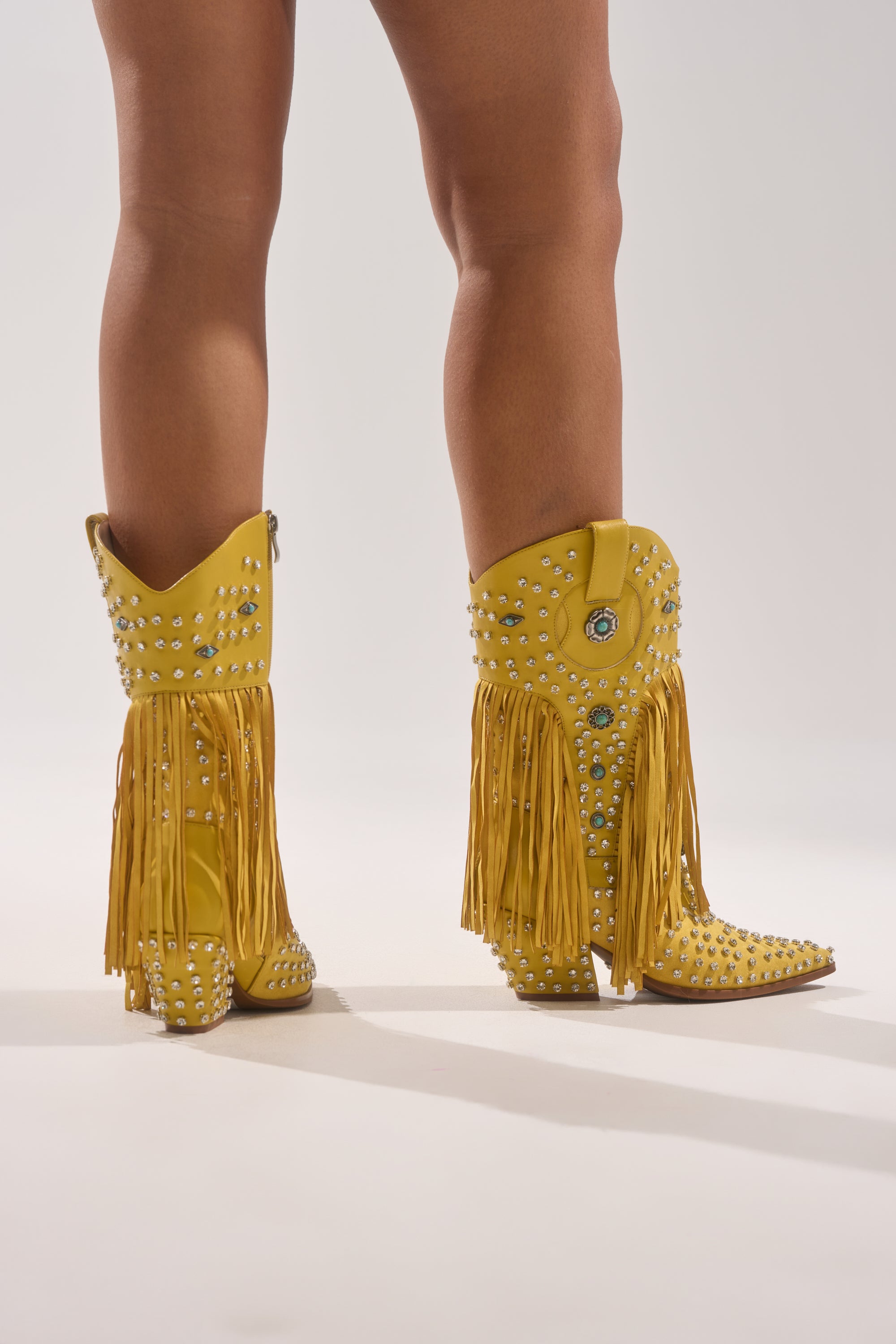 A person stands on a plain light background wearing the AZALEA WANG GREYSON YELLOW RHINESTONE EMBELLISHED WESTERN BOOTIE featuring yellow faux leather, silver studs, long fringe, rhinestone details, and pointed toes. Legs are visible from knee down.