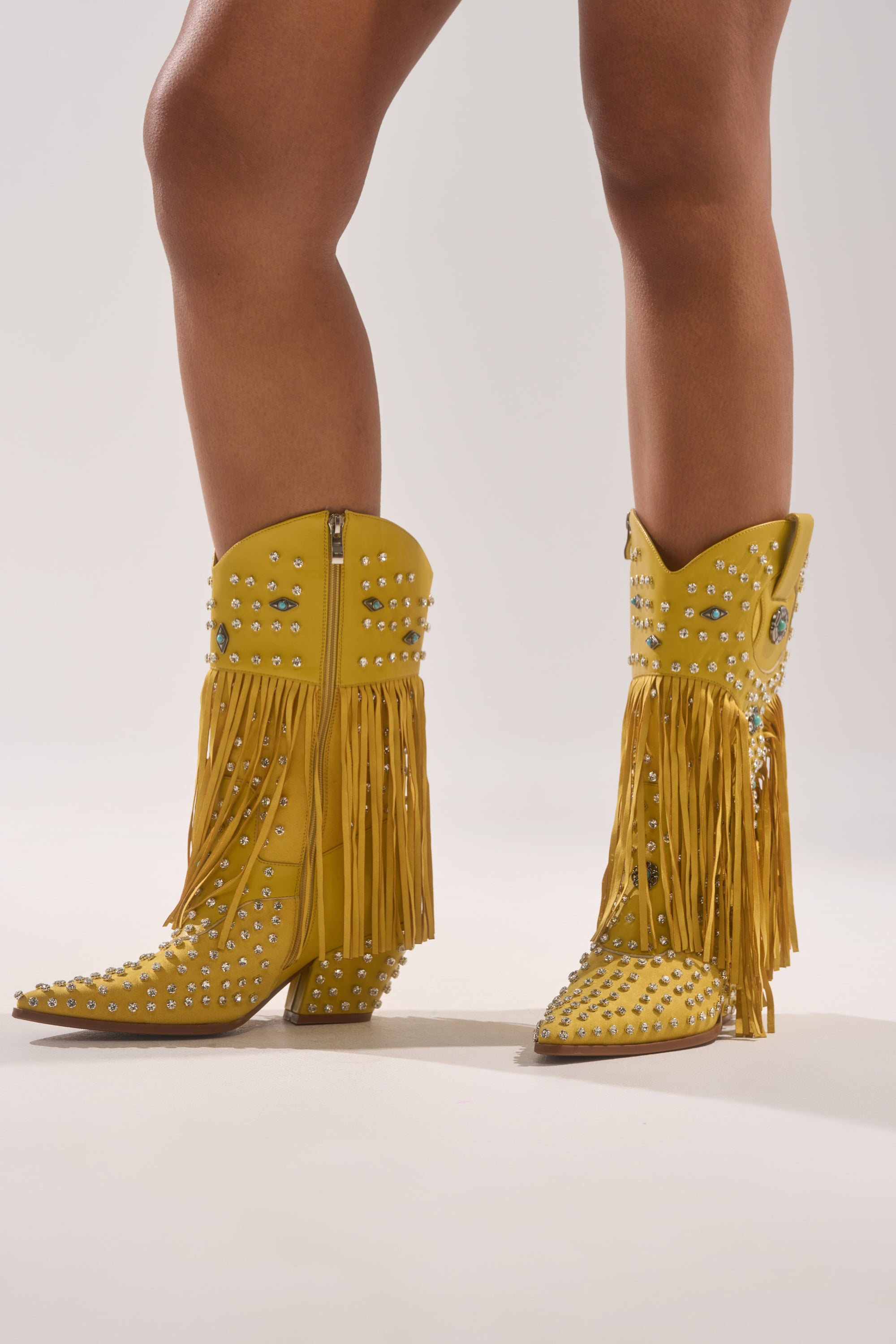 Someone wears the AZALEA WANG GREYSON YELLOW RHINESTONE EMBELLISHED WESTERN BOOTIE, featuring turquoise accents and long fringe, standing against a plain light background.