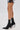 A person wearing a geometric black and white skirt and AZALEA WANG GRYFFON BLACK STILETTO BOOTIE—faux leather booties with side zippers and rainbow spike-studded heels—stands against a plain white background.