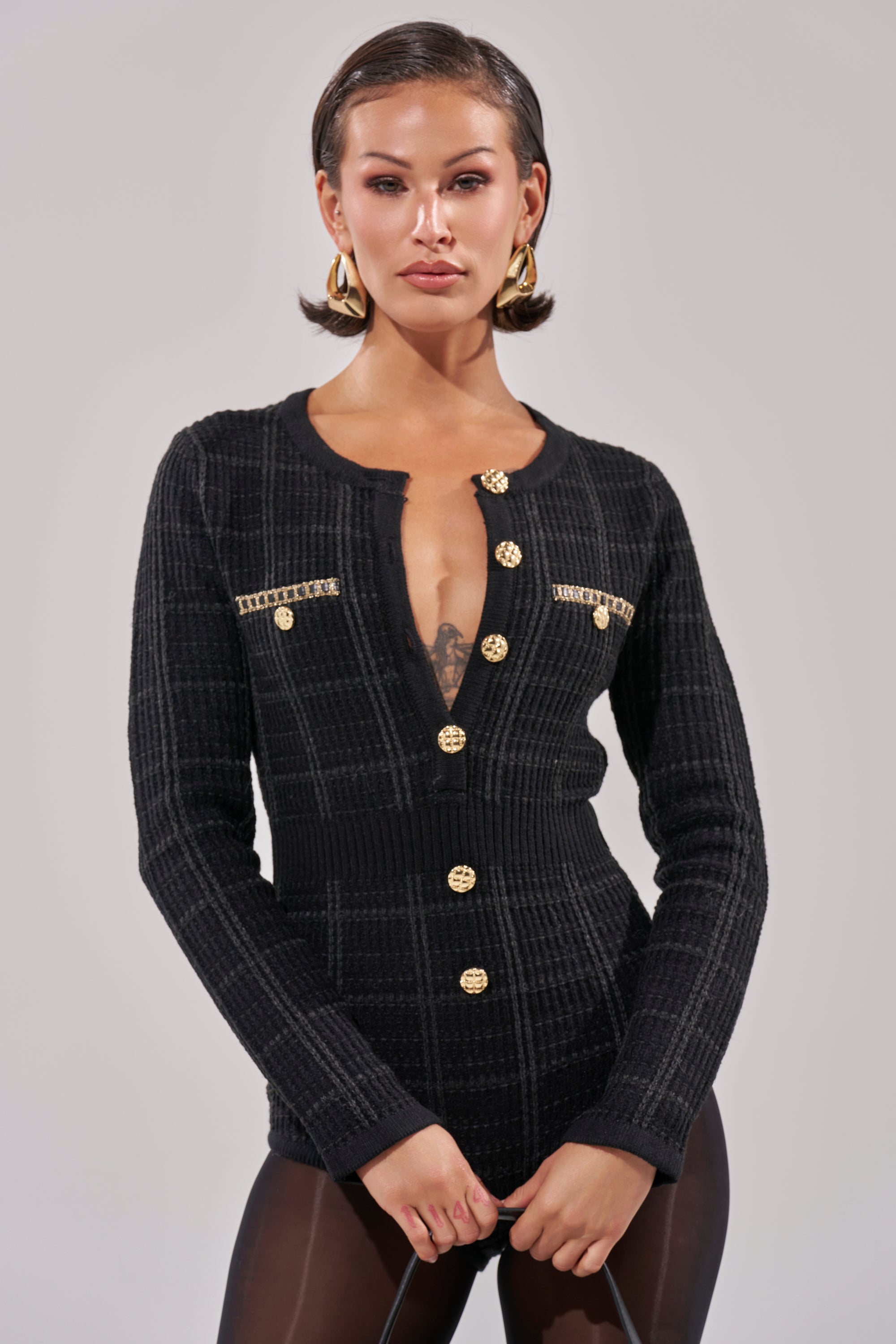 A woman with short, slicked-back hair wears the HOLIDAY TIME PLAID ROMPER with gold buttons and hoop earrings, standing with hands clasped. Her look exudes confidence and chic holiday style against a plain background.