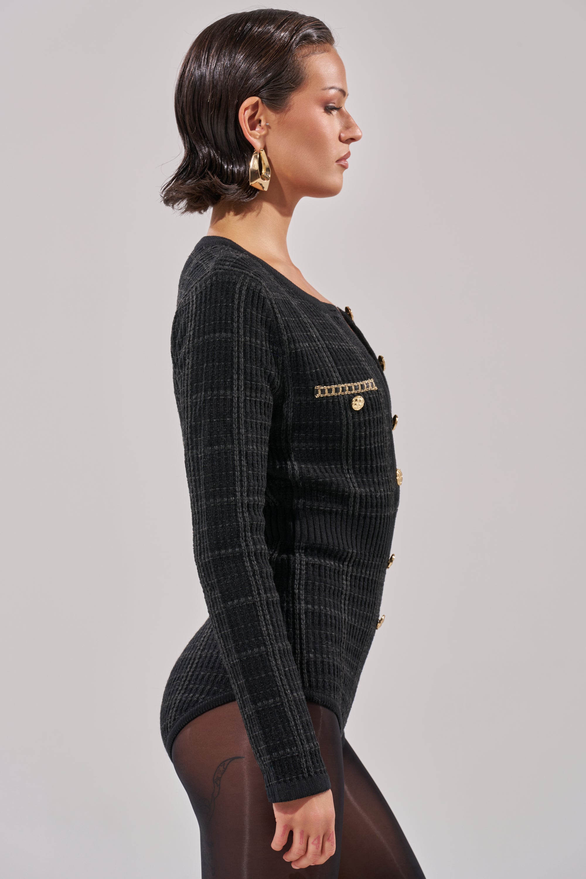 A woman with short slicked-back hair, gold earrings, and a fitted black top poses in profile against a plain light background—perfect inspiration for styling the HOLIDAY TIME PLAID ROMPER this holiday season.