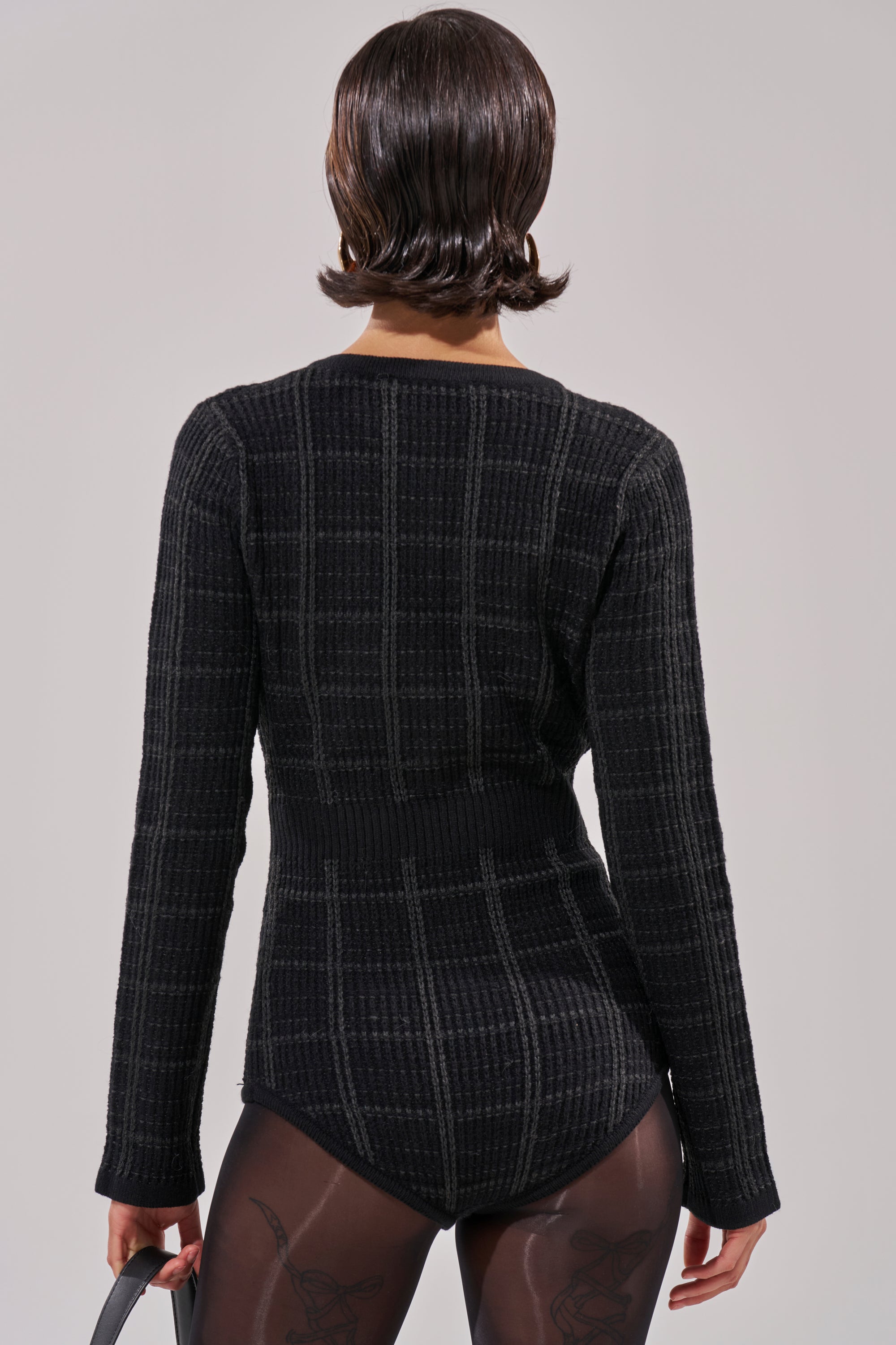 A person with short, dark, wavy hair is seen from behind in a textured black bodysuit and sheer black tights, perfect for layering under the HOLIDAY TIME PLAID ROMPER and accessorized with a gray bag in their left hand.