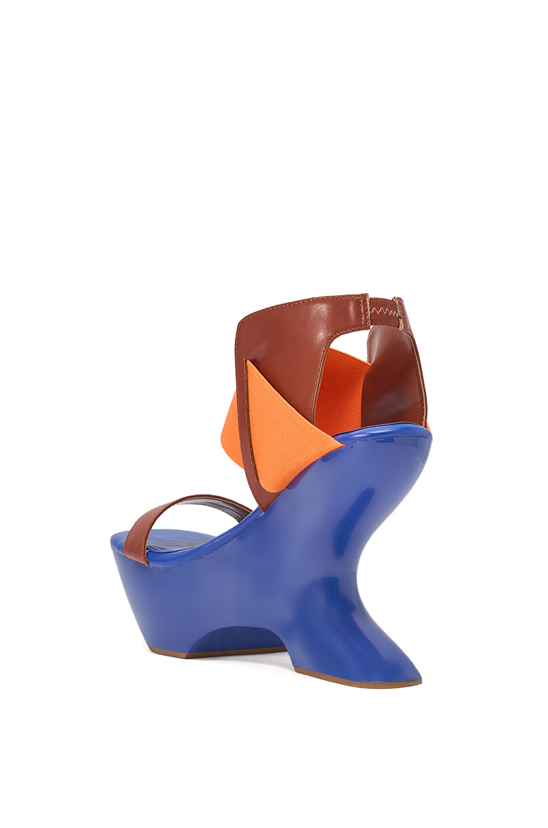 The AKIRA GOLD GUILLAUME BLUE MULTI NOVELTY COLOR BLOCK WEDGE SANDAL features a sculptural high heel, glossy blue platform, brown faux leather ankle strap, orange fabric accent, and open toe, shown from the rear against a white background. 4