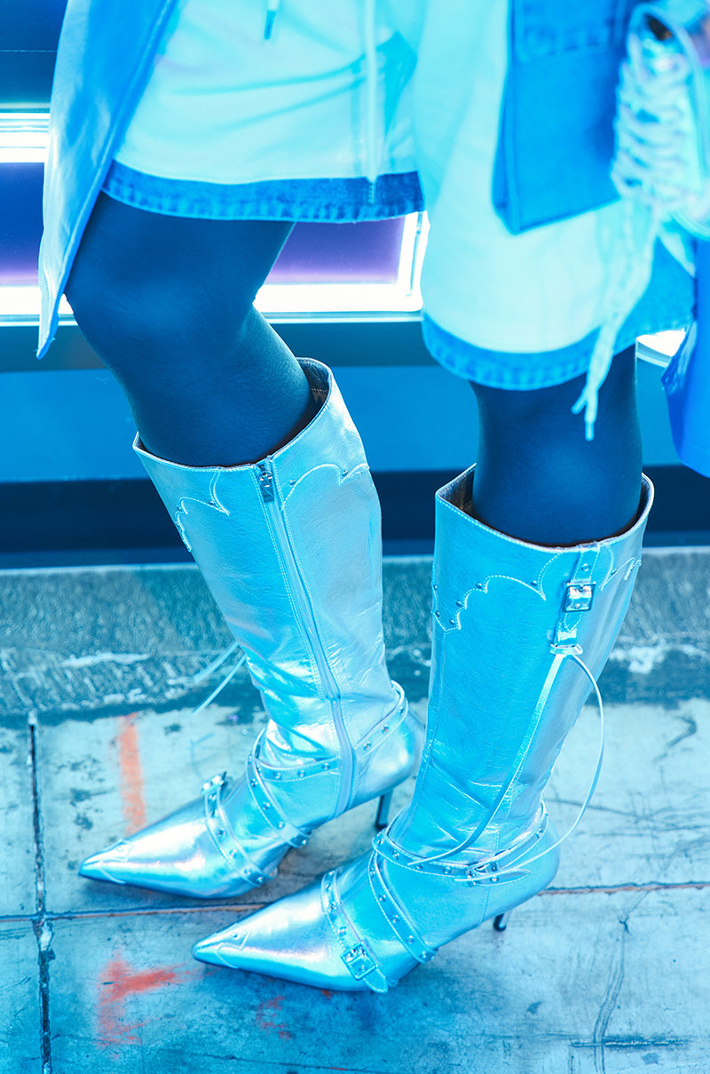 A person wearing the GUNNER SILVER STILETTO BOOT, a metallic faux leather knee-high design featuring pointed toes and buckles, is paired with denim shorts and a long coat. The blue lighting casts a futuristic glow on the scene, while orange markings decorate the floor.