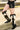 A person stands outdoors near a yellow metal railing, wearing black denim shorts and the AZALEA WANG GWAYNE BLACK MULTI COW WESTERN BOOT, with one leg crossed over the other.