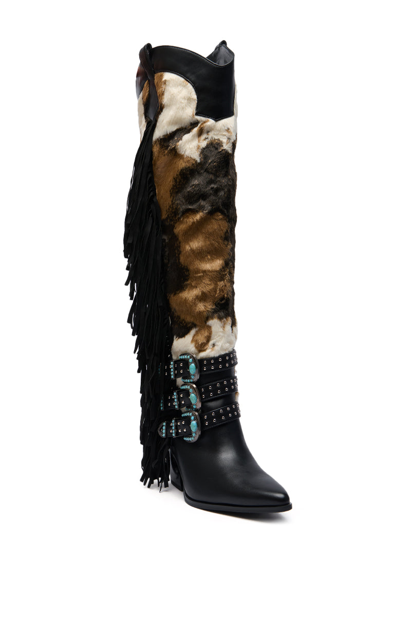 The AZALEA WANG GWAYNE BLACK MULTI COW WESTERN BOOT features knee-high faux fur, a pointed toe, black leather accents, a brown and white cow print, black fringe, and three black straps with turquoise buckles and studs.