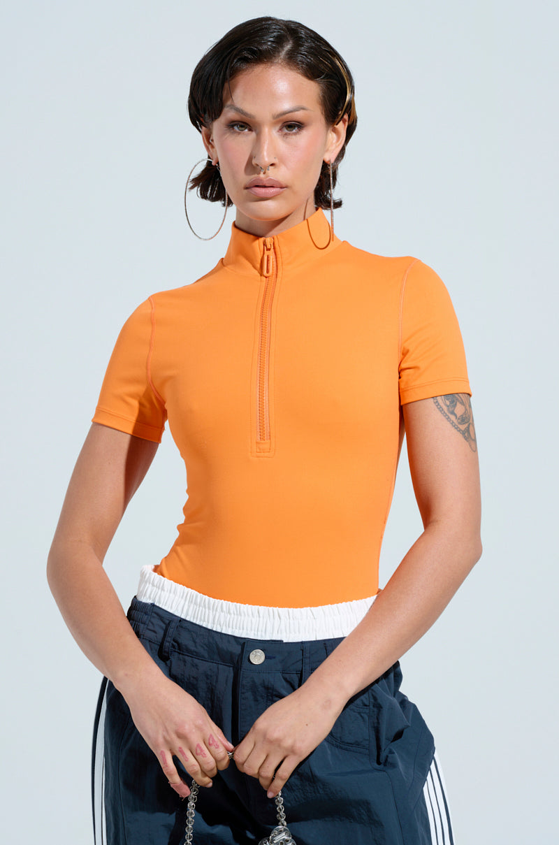 Confident in a PAXTON ZIP UP SHORT SLEEVE BODYSUIT, a person with short dark hair and large hoop earrings holds a bag. They pair the orange bodysuit stylishly with navy pants and a white waistband, displaying a tattoo on their right arm against a plain background.