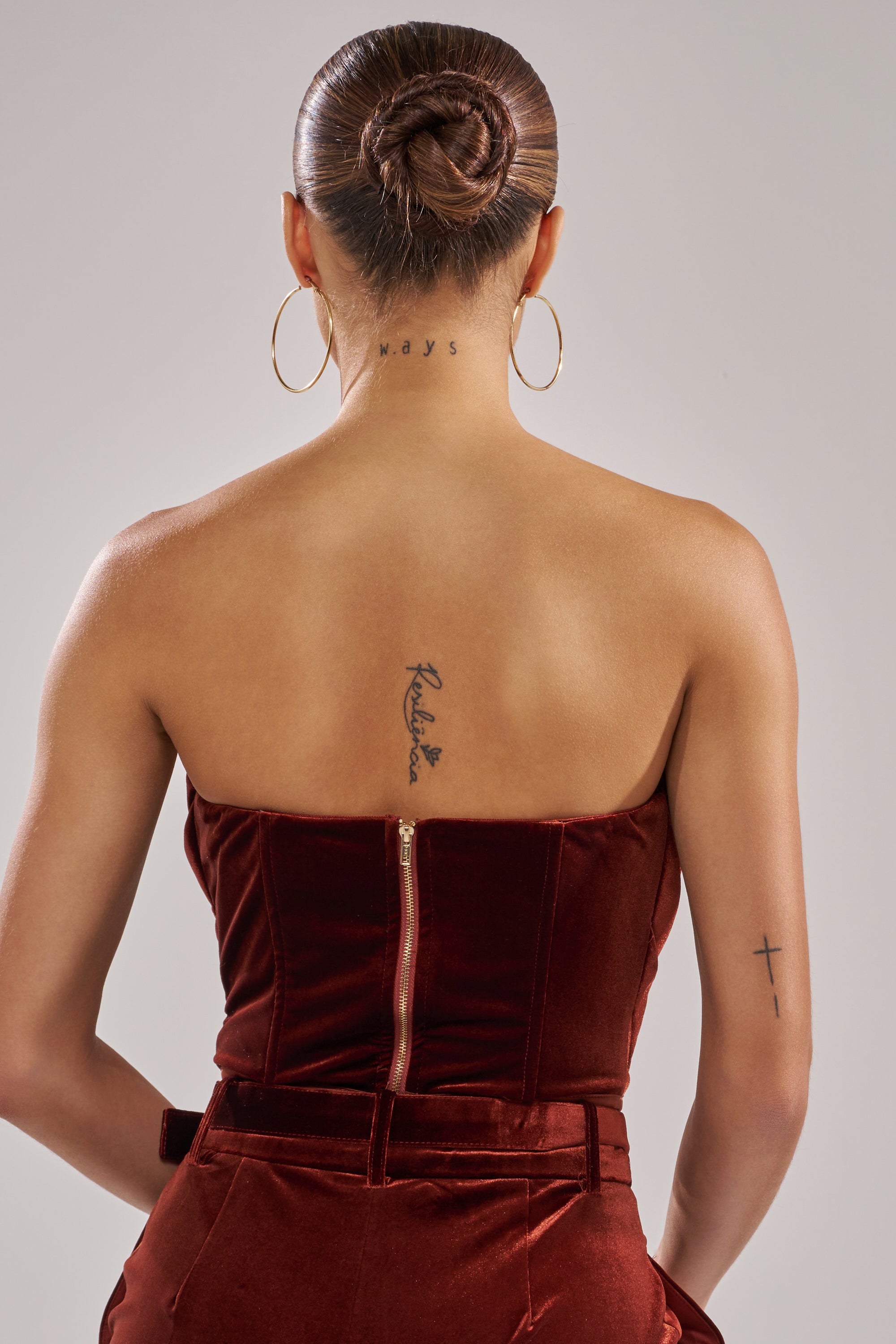 A woman wearing large hoop earrings and the FLASHING LIGHTS VELVET CORSET, styled in a neat bun, is shown from behind. Visible tattoos include text on her neck, script on her upper back, and a cross on her arm.