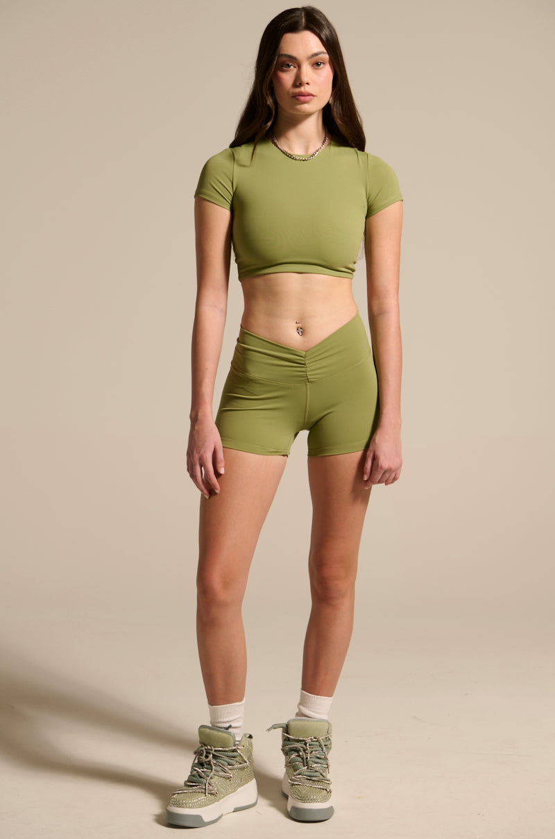 A young woman stands facing forward in PAXTON SEXY RUCHED BIKER SHORTS and a fitted light green crop top, styled with light socks and chunky sneakers in a neutral studio setting.