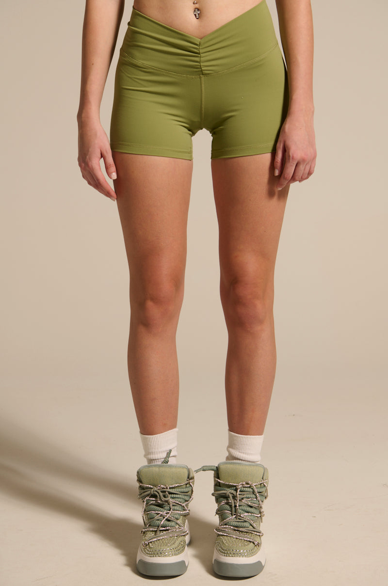 A person wears buttery soft, high-rise fit PAXTON SEXY RUCHED BIKER SHORTS in light green, paired with white socks and light green lace-up boots, standing against a plain beige background. The person's torso is partially visible.