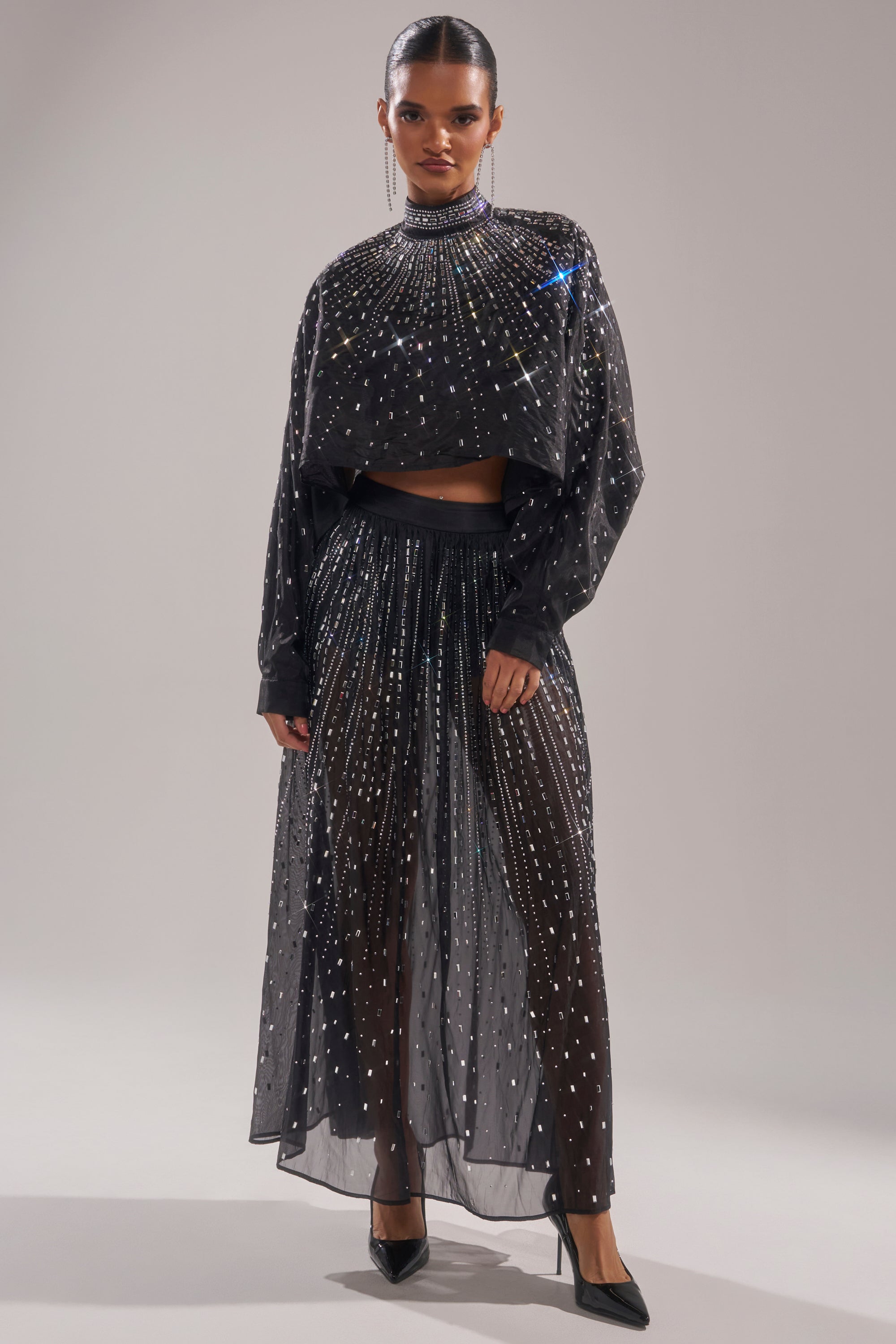 A woman poses in the DANCING QUEEN RHINESTONE MESH MAXI SKIRT, a black sheer outfit with sparkling rhinestones, styled with a loose long-sleeve top over black shorts and finished with black pointed heels. 0