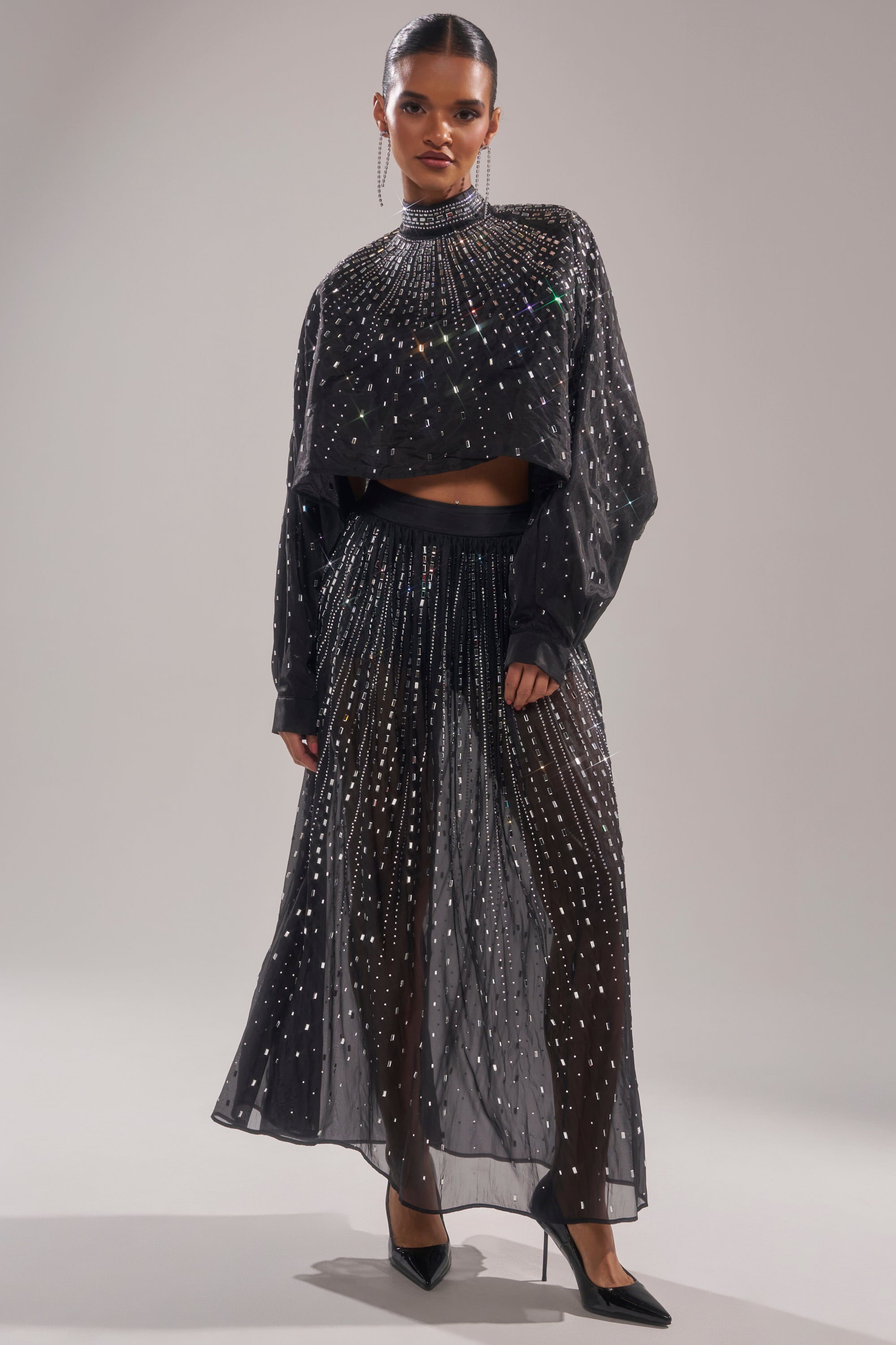 A woman poses in the DANCING QUEEN RHINESTONE MESH MAXI SKIRT and a matching long-sleeve top, both sheer and adorned with sparkling rhinestones. She completes the look with black heels, drop earrings, and slicked-back hair against a light background. 6