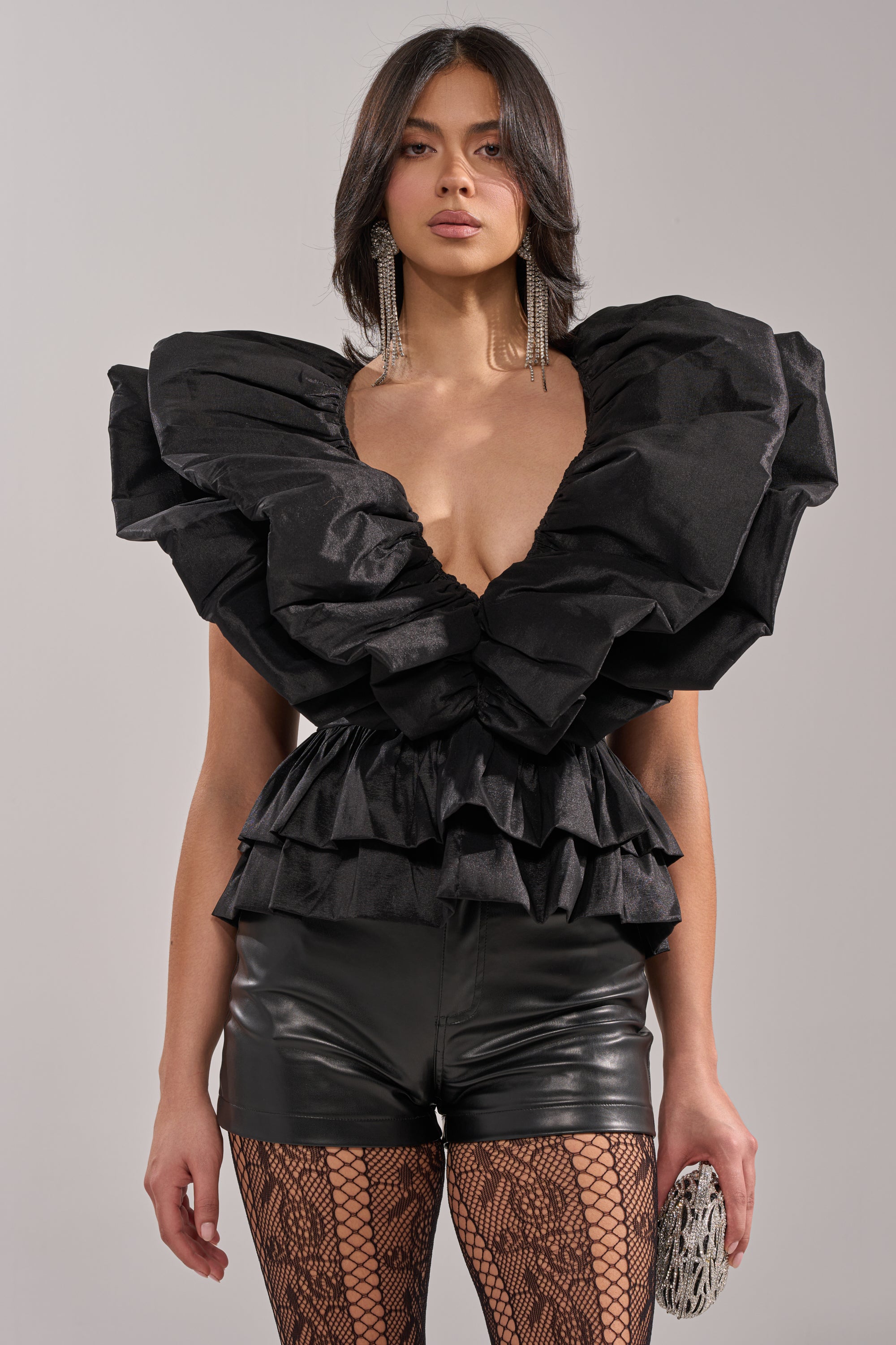 A person stylishly poses in the WORK OF ART BLOUSE—a sculptural black ruffled organza top with a deep V-neck—paired with black leather shorts, fishnet stockings, dangling earrings, and a silver clutch, creating perfect statement eveningwear. 1