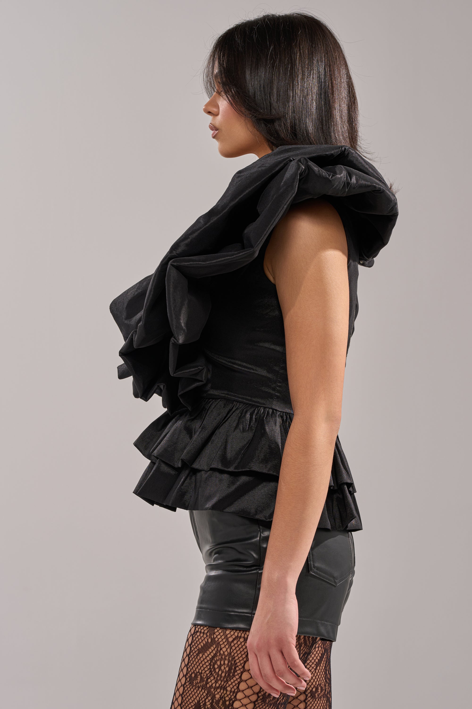 A woman in profile models the WORK OF ART BLOUSE— a black, ruffled organza top with dramatic layers—paired with black leather shorts and fishnet stockings. She stands before a plain gray background, her dark hair straight and shoulder-length. 2