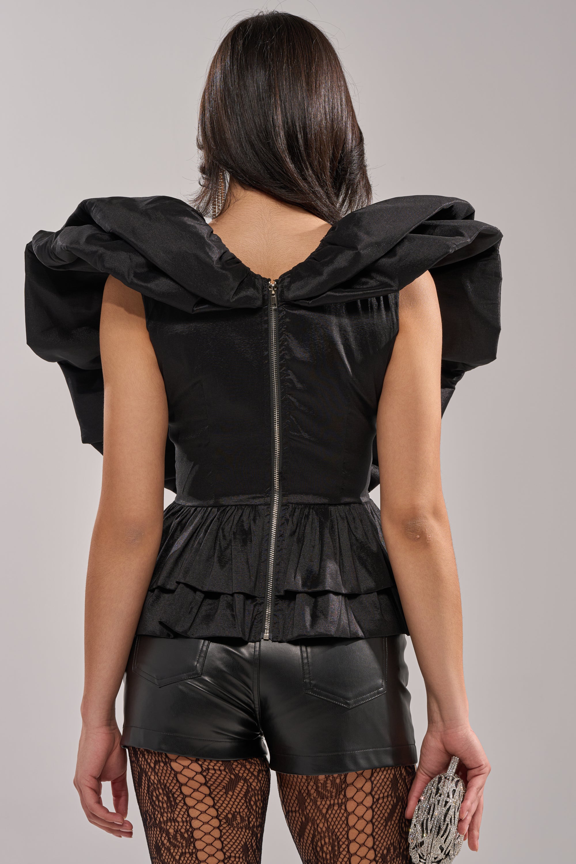 A woman with long dark hair stands facing away, wearing the WORK OF ART BLOUSE with dramatic ruffled organza shoulders, paired with black leather shorts, fishnet tights, and a small textured clutch—a true statement look. 3