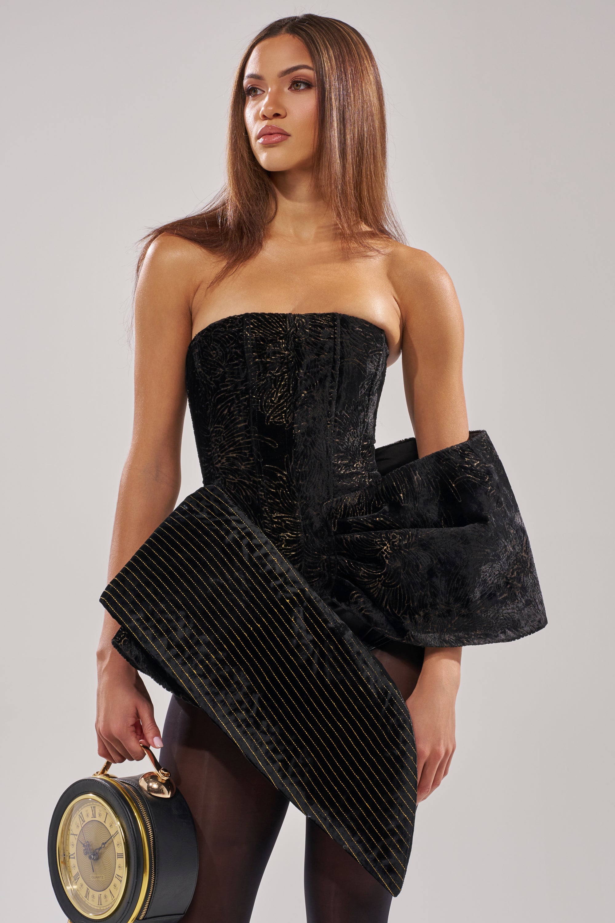 A woman wears the DRAMA CORSET IN BLACK, a strapless asymmetrical dress with gold embroidery. She holds a clock-shaped handbag, has long straight hair, and wears sheer black tights against a plain light background.