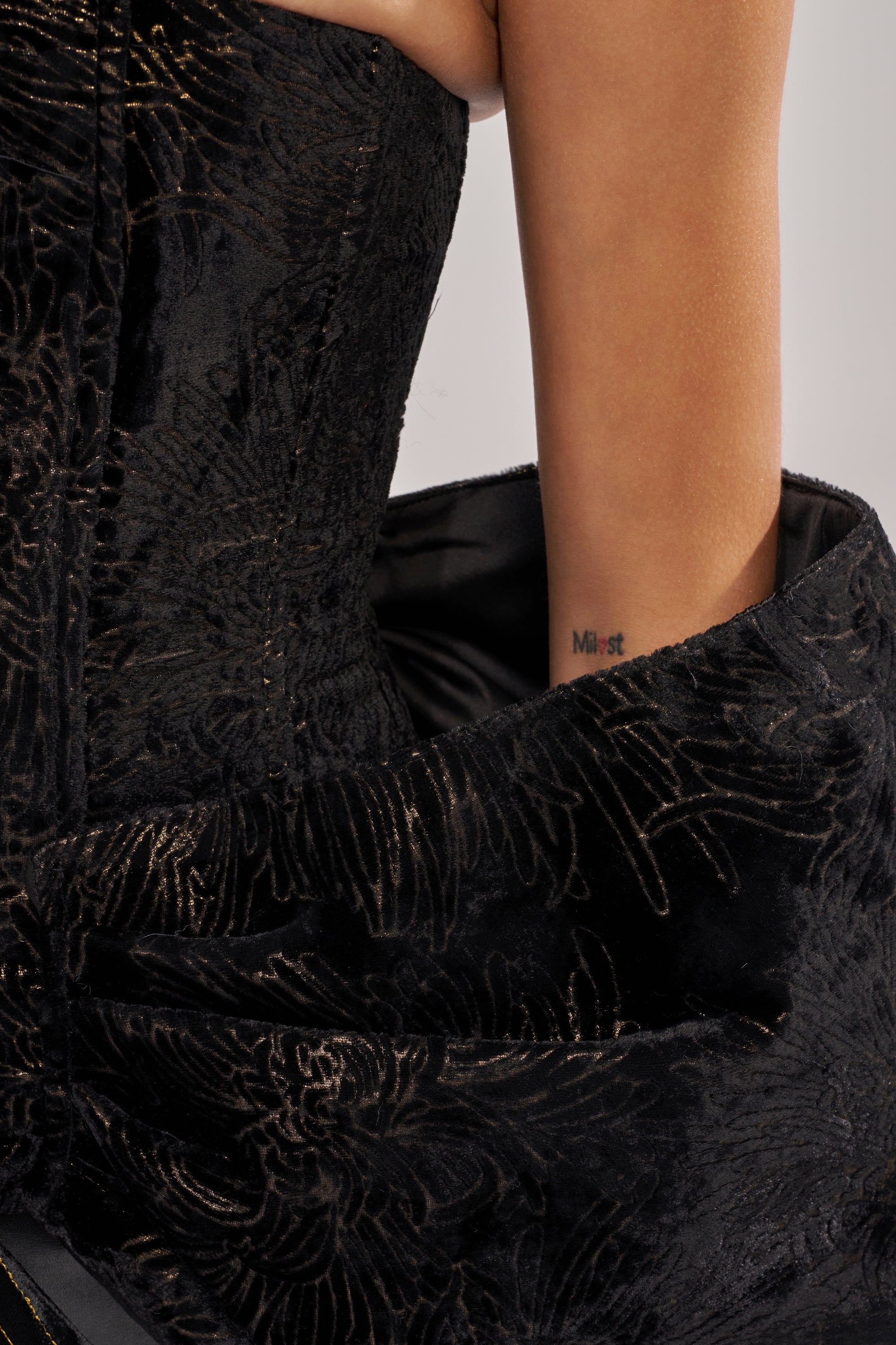 A close-up of a person in the DRAMA CORSET IN BLACK, a textured velvet dress with gold floral embroidery, revealing the side of their bare arm and an inner arm tattoo that reads “MIST.”.