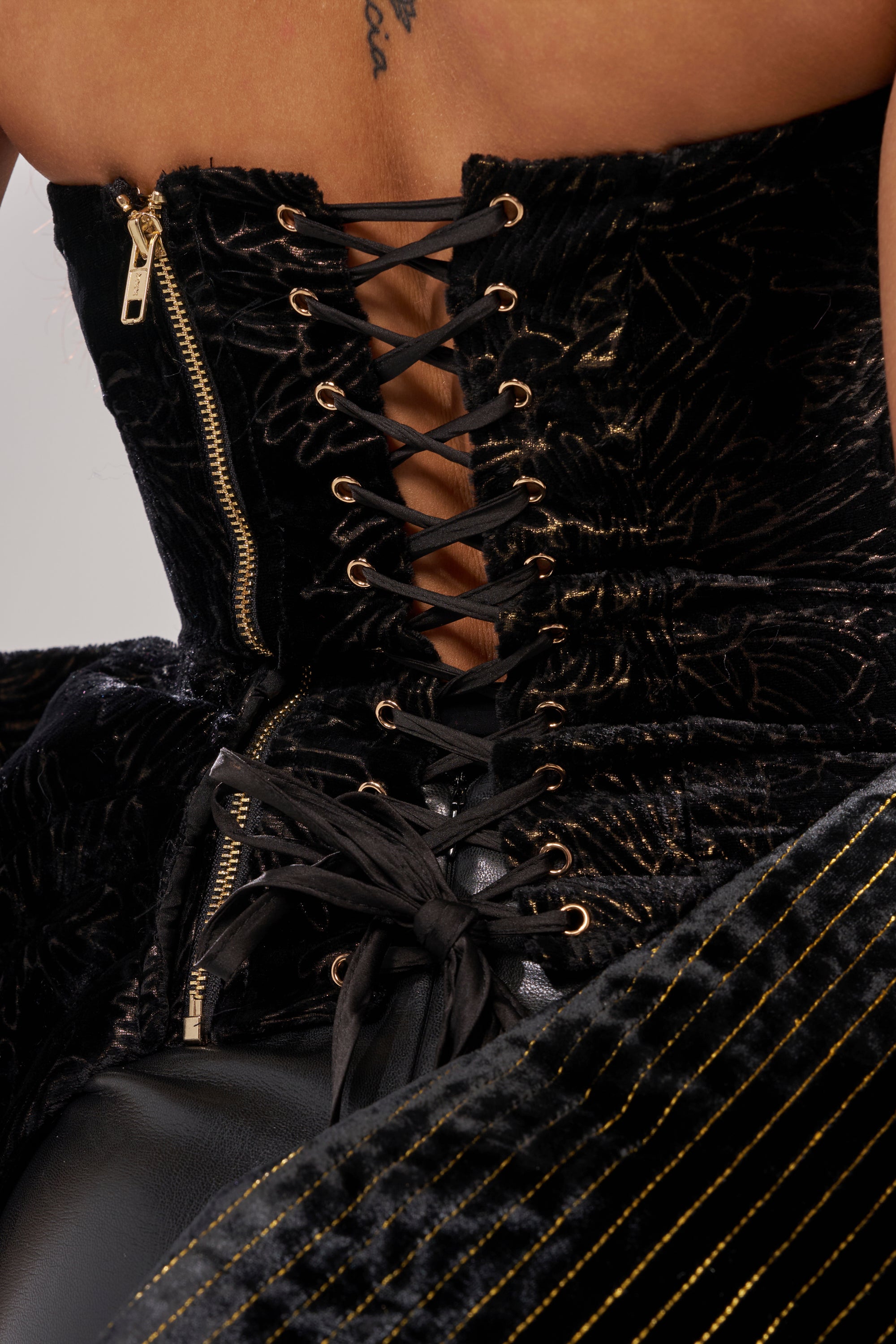 Close-up of the DRAMA CORSET IN BLACK, featuring black velvet, gold eyelets, a gold zipper, black laces tied in a bow, subtle patterns with gold embroidery details, and thin gold stripes adding dramatic contrast.