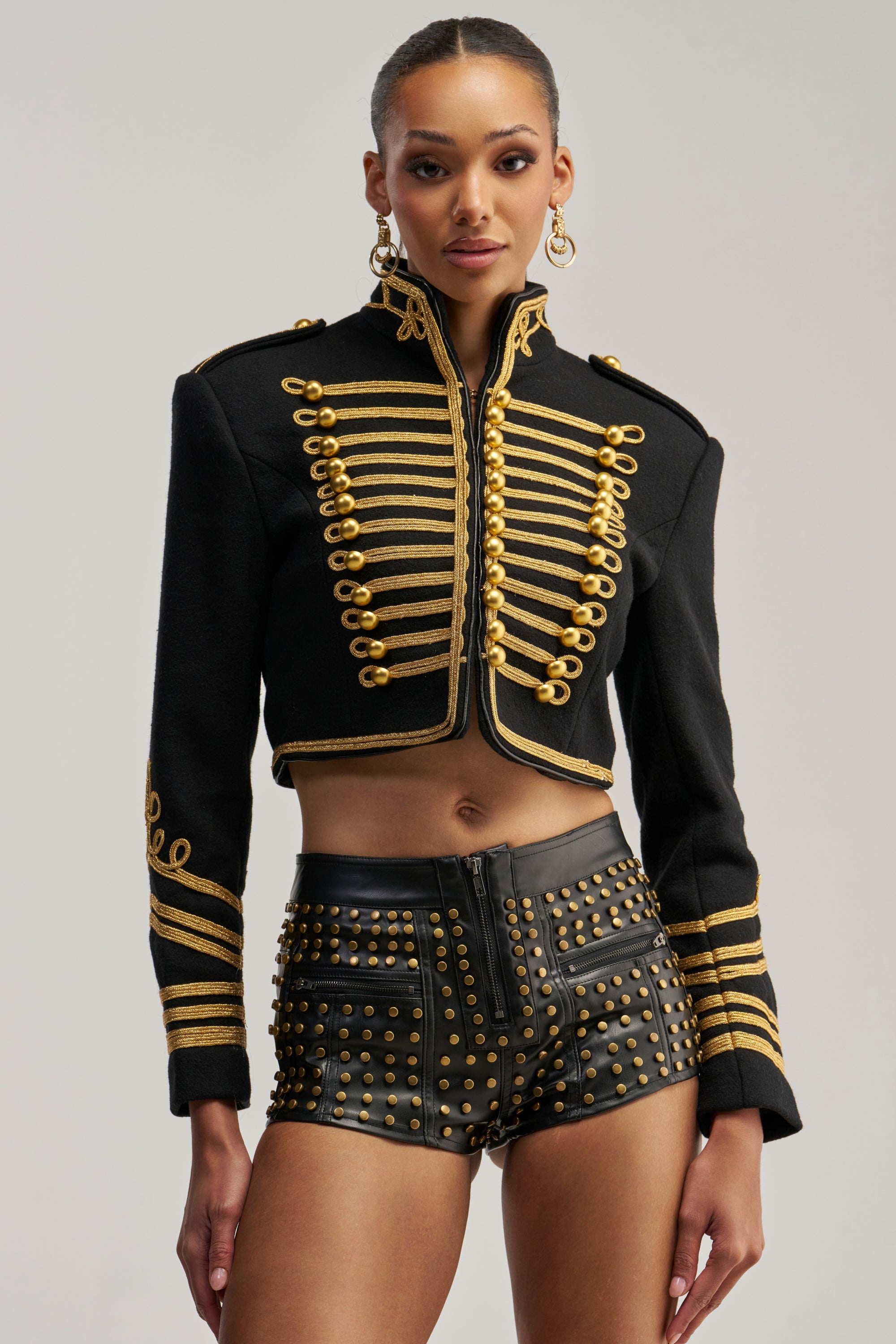 Wearing the VIVIANNE MILITARY JACKET—a black, cropped military-inspired piece detailed with gold accents and buttons—paired with high-waisted black leather shorts embellished with gold studs.