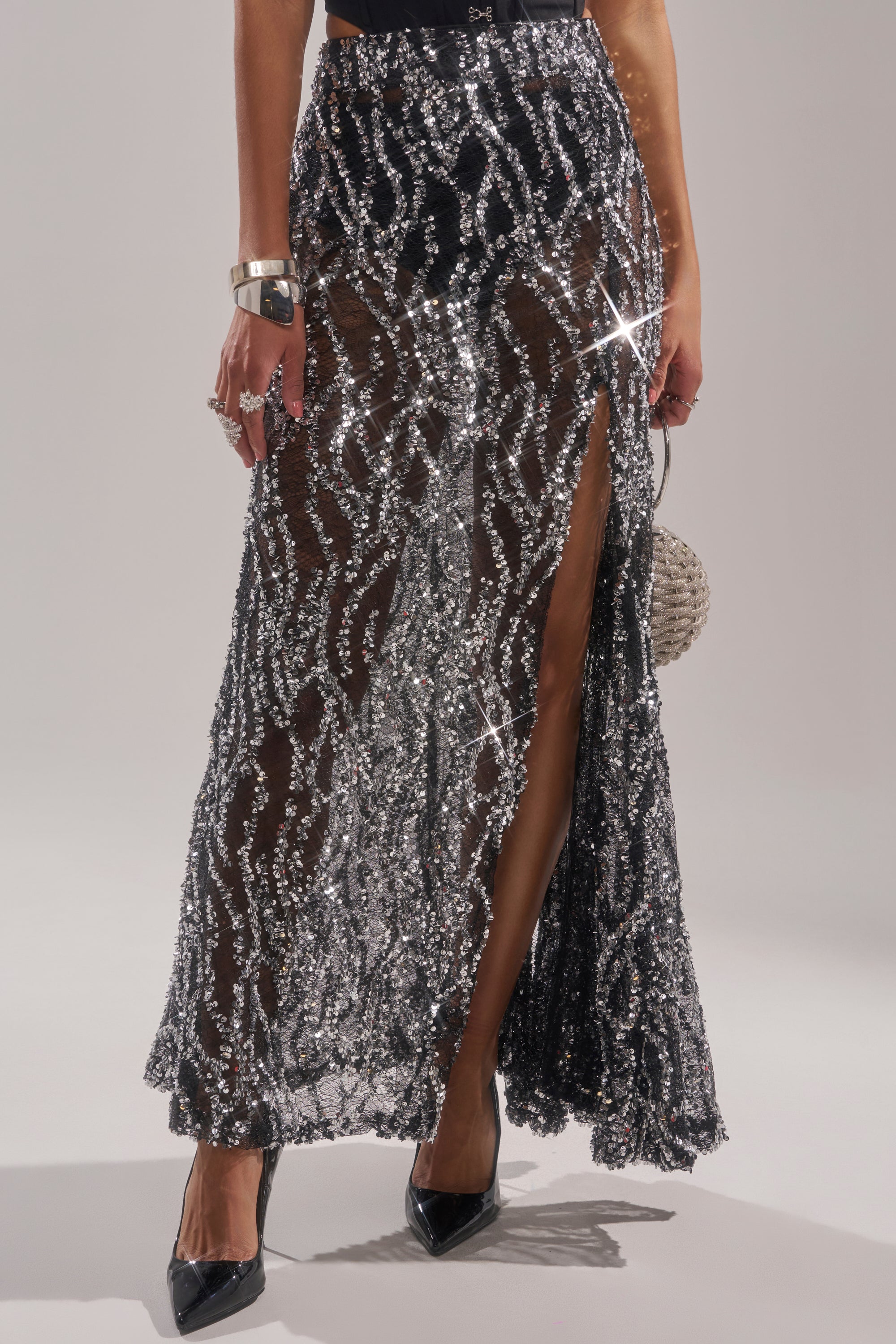 Wearing the BEAUTIFUL SEQUIN MAXI SKIRT with a thigh-high slit, black heels, silver jewelry, and a round clutch bag. The sparkling silver skirt catches the light for a dazzling effect. 1