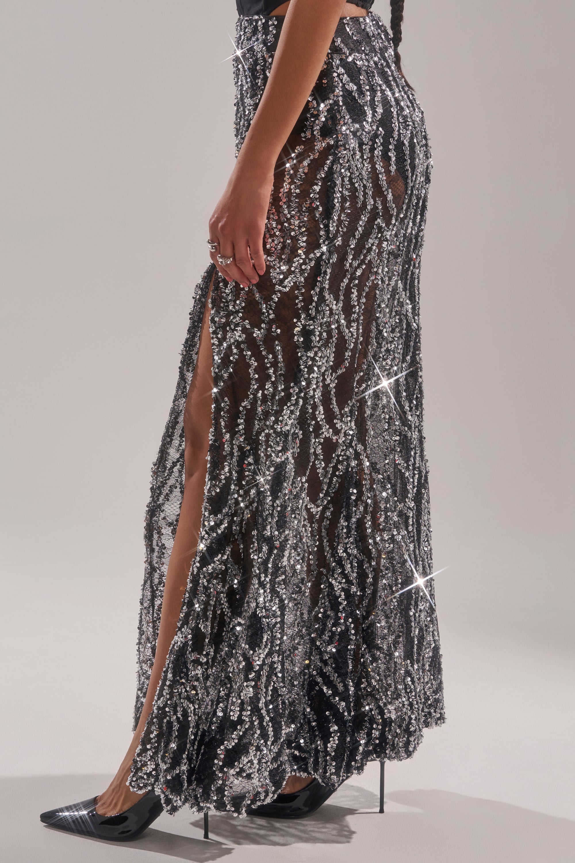 A person wearing the BEAUTIFUL SEQUIN MAXI SKIRT, a sheer maxi embellished with silver sequins in swirling patterns and a thigh-high slit, paired with black pointed-toe heels. 3
