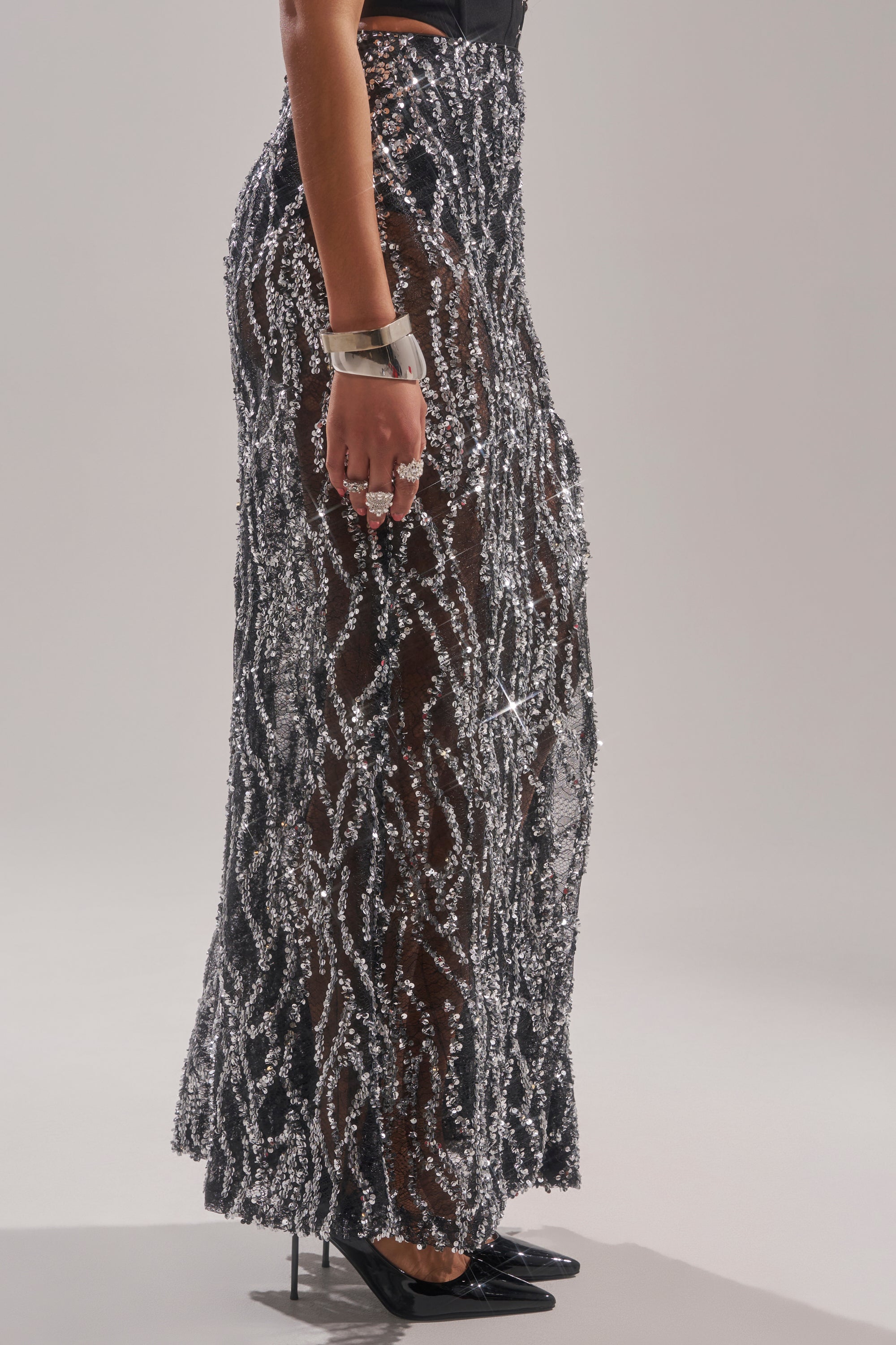 Wearing the BEAUTIFUL SEQUIN MAXI SKIRT adorned with silver beads, paired with black pointed heels and silver jewelry, a person stands against a plain light background. 4
