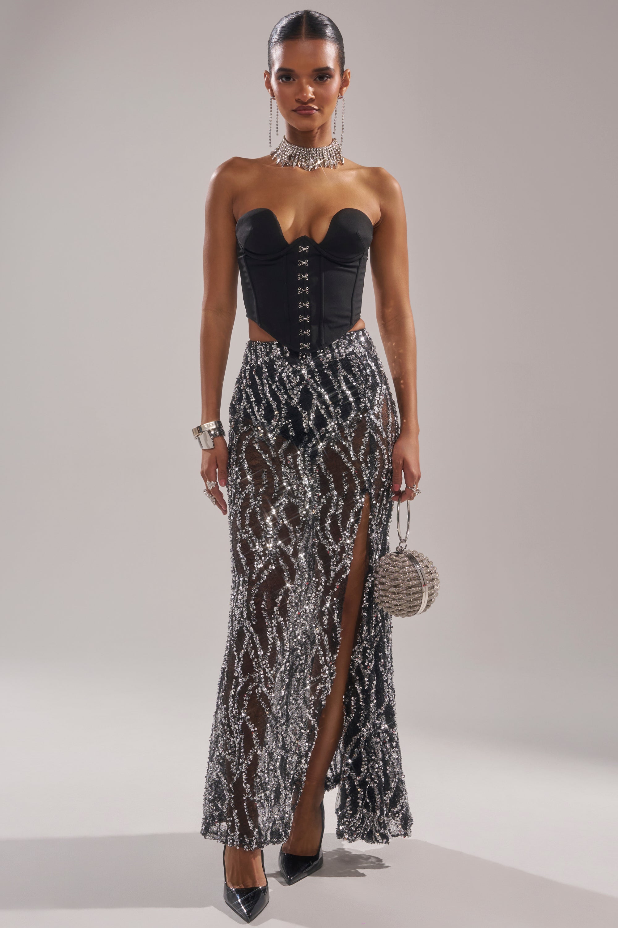 A woman wears the BEAUTIFUL SEQUIN MAXI SKIRT, a sheer silver sequin skirt with a high slit, paired with a strapless black corset top. She accessorizes with a statement necklace, bracelets, a round silver handbag, and black heels. 6