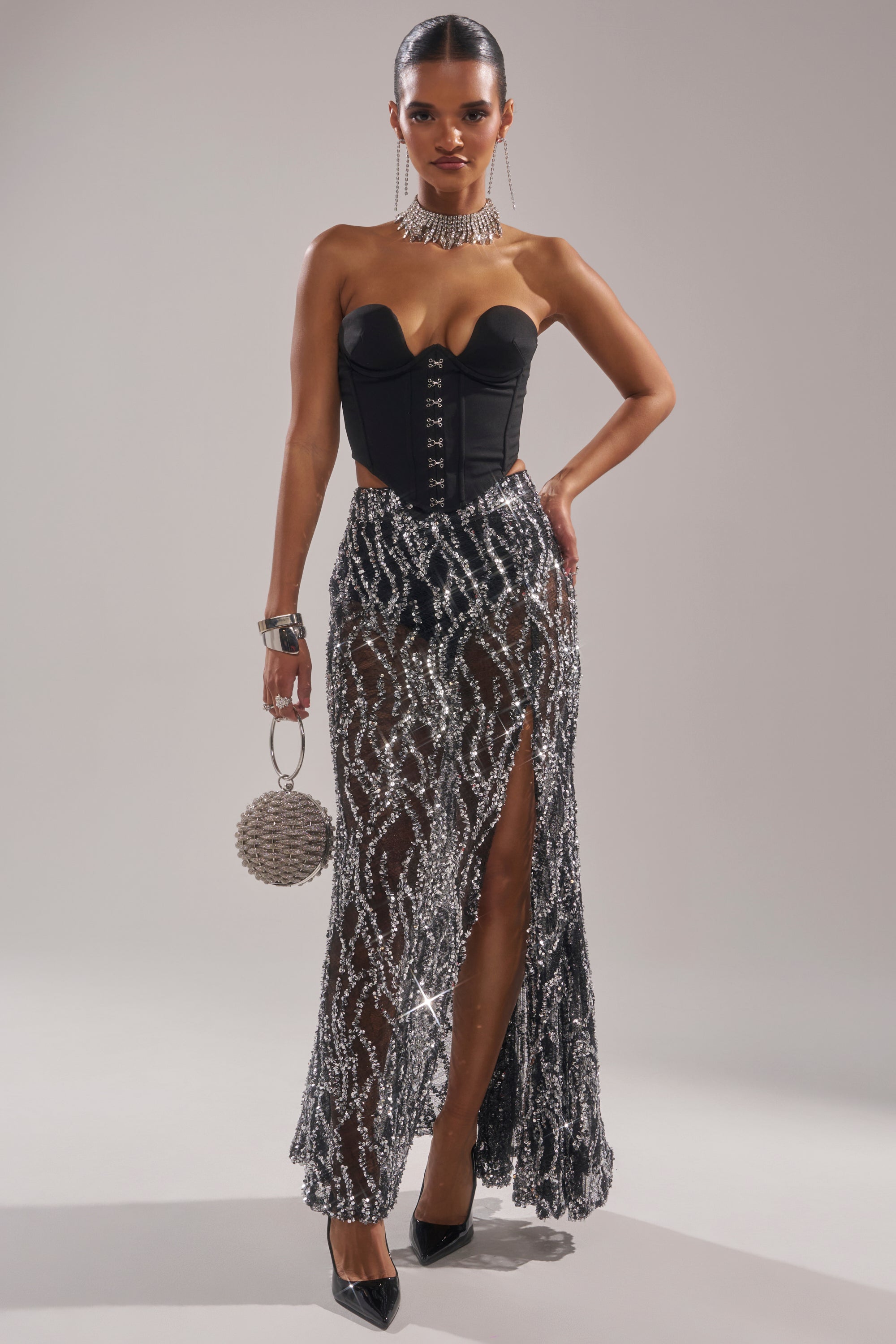 A woman poses confidently in a strapless black corset top and the BEAUTIFUL SEQUIN MAXI SKIRT, accessorized with a statement necklace, round beaded purse, and black heels against a neutral backdrop. 7