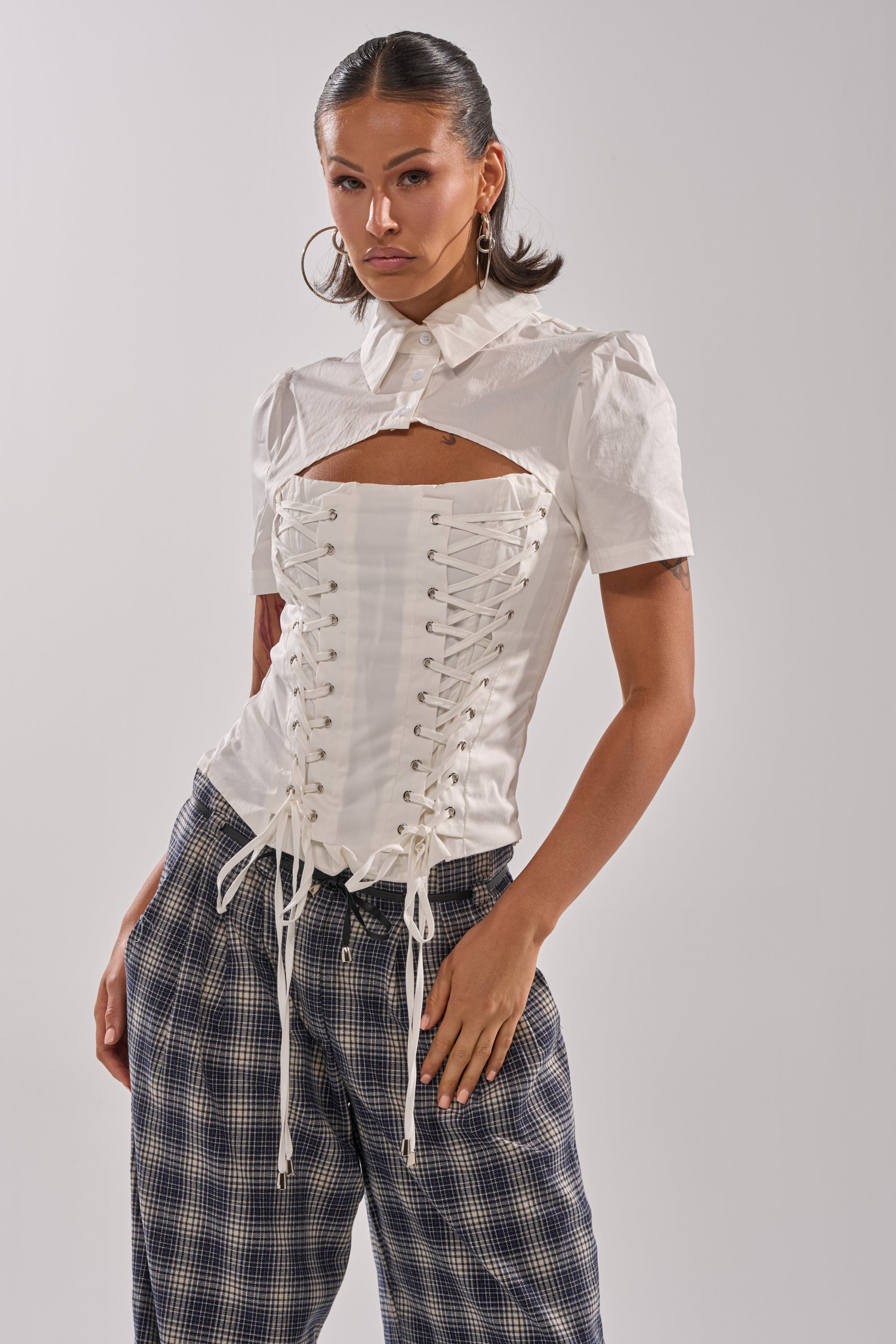 A woman models the BUSINESS ONLY BLOUSE, a white cotton-blend top with lace-up and cutout front details, paired with loose plaid pants. She accessorizes with large hoop earrings and has her hair slicked back.