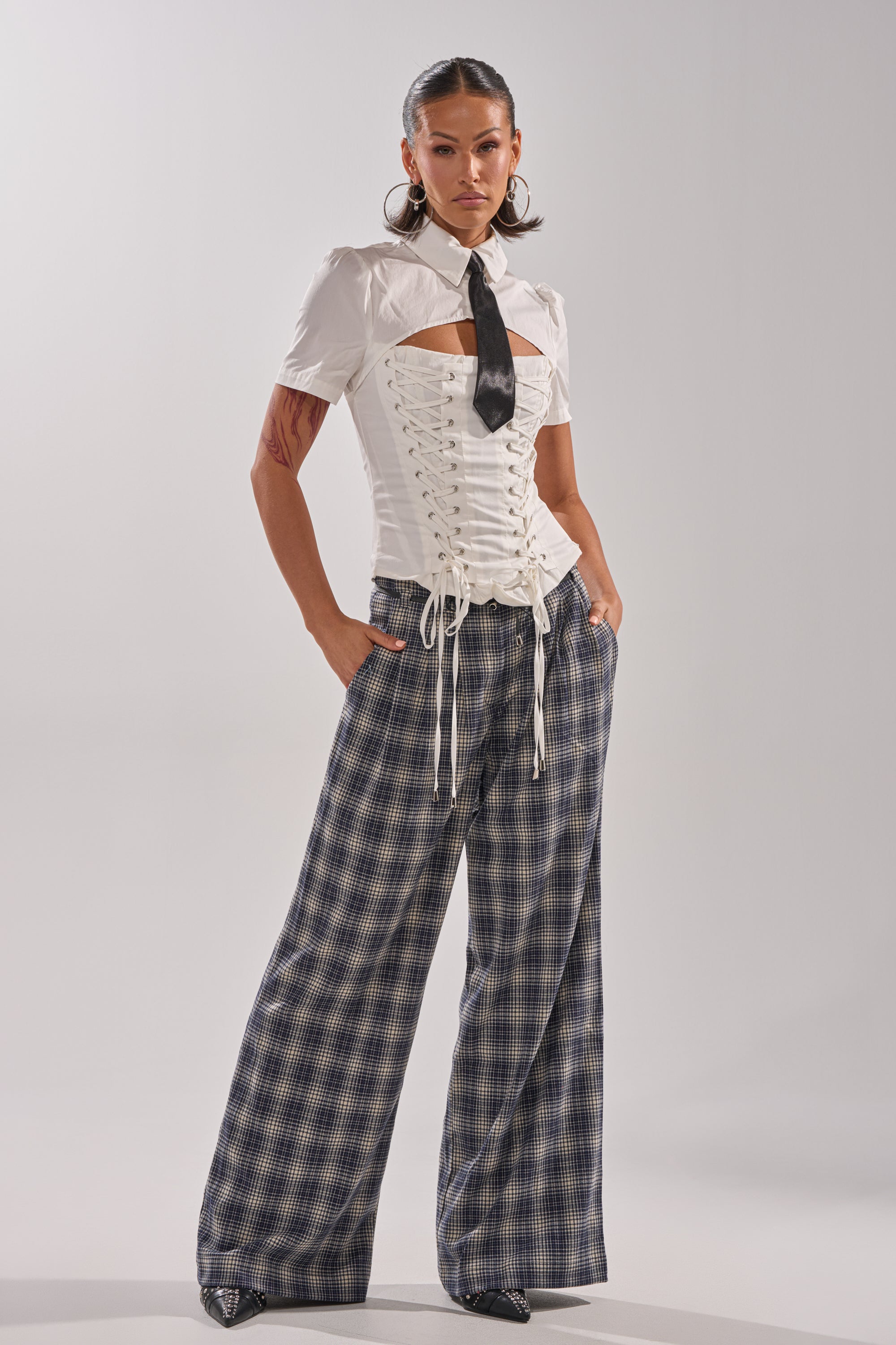 A woman stands confidently wearing the BUSINESS ONLY BLOUSE—a short-sleeved cotton blend with a lace-up corset overlay—paired with a black tie, wide-leg plaid pants, and pointed shoes against a plain light background.