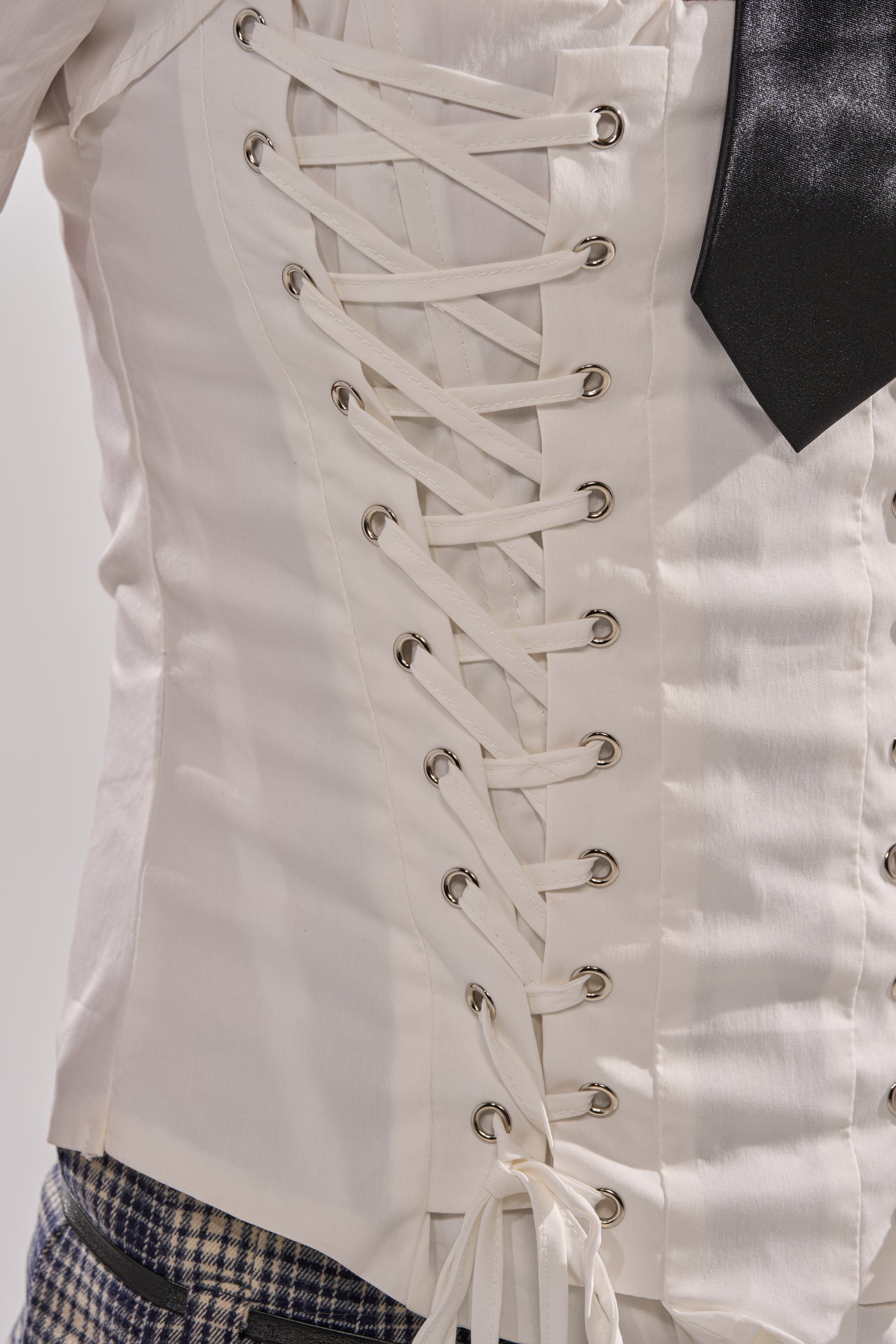 Close-up of the BUSINESS ONLY BLOUSE, a white lace-up corset with silver eyelets and white ribbon detail, styled with a black tie and checkered pants.