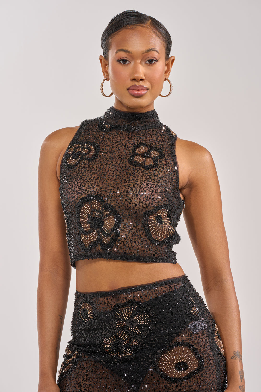 A woman stands against a plain background wearing the ALL EYES ON ME FLORAL MOCKNECK TOP—sleeveless, sheer, with sequin floral patterns. She pairs it with a matching skirt, hoop earrings, and short hair for a confident evening party look. 1