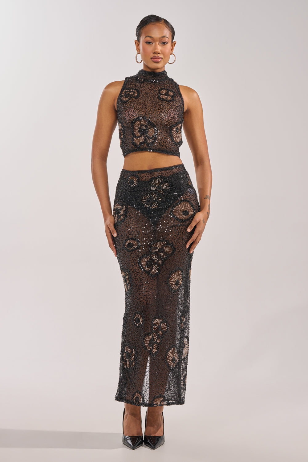 A woman faces forward in the ALL EYES ON ME FLORAL MOCKNECK TOP, a black sheer sequin crop top with floral patterns, paired with a matching skirt, hoop earrings, slicked-back hair, and black pointed heels—an elegant party ensemble. 5
