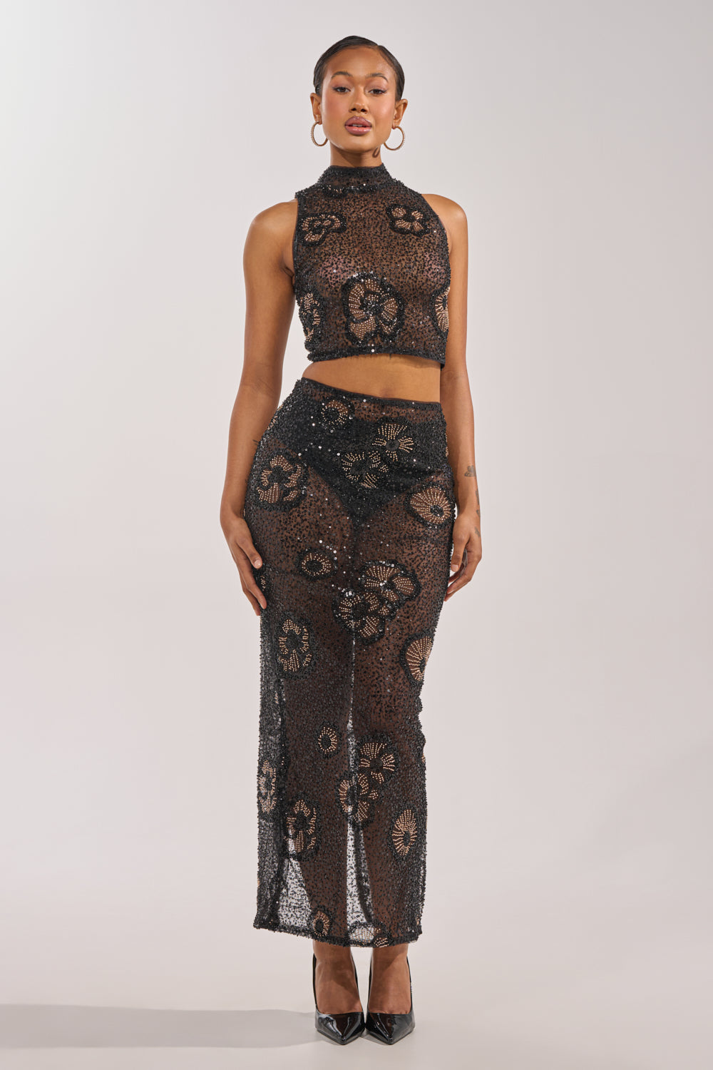 A woman faces forward in the ALL EYES ON ME FLORAL MOCKNECK TOP, a sleeveless, sheer floral piece paired with a matching long skirt. She accessorizes with hoop earrings, slicked-back hair, and black heels against a light background. 6