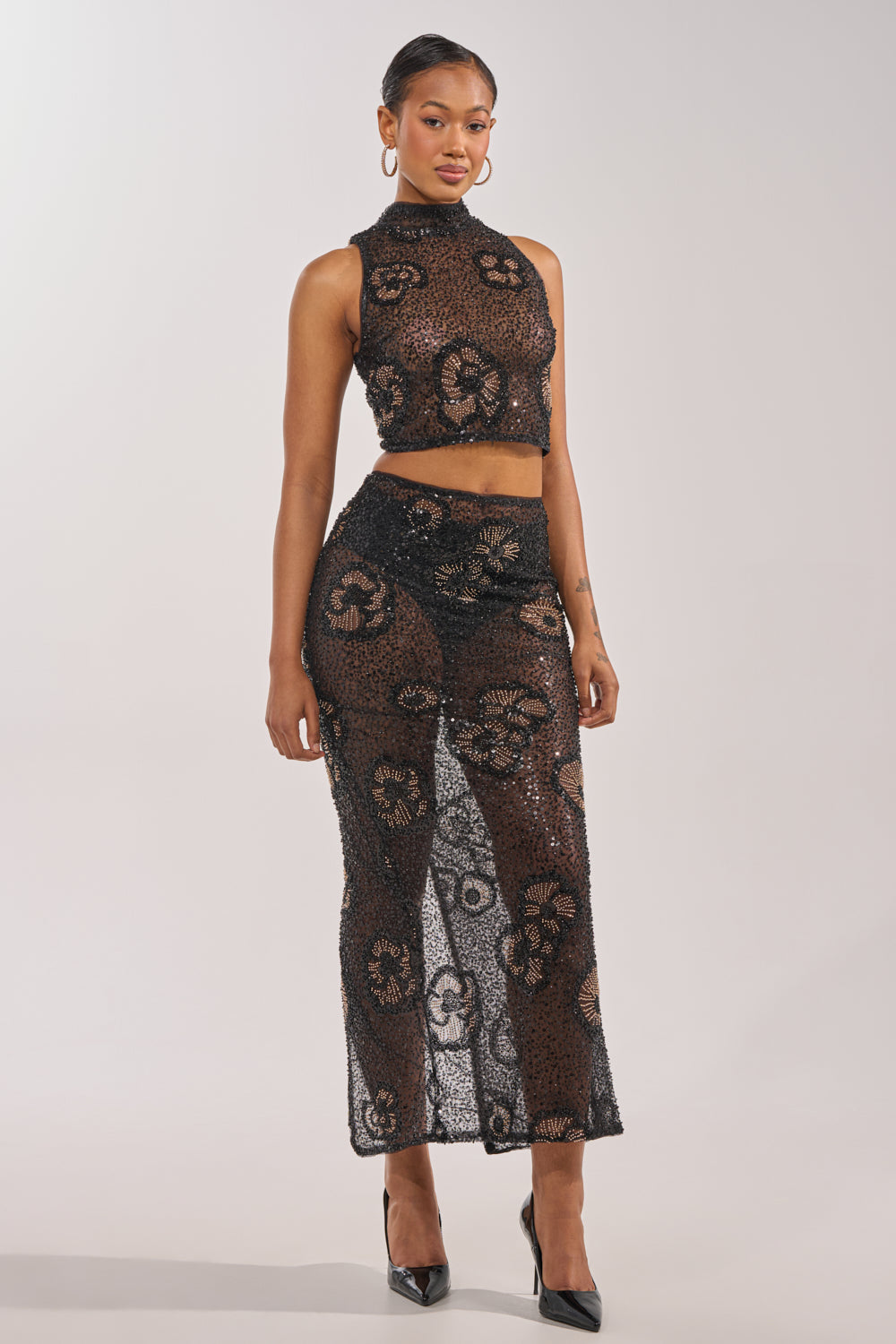 A woman faces forward in the ALL EYES ON ME FLORAL MOCKNECK TOP, a black beaded embroidered piece paired with a sheer, patterned skirt featuring circular floral motifs and black heels. The scene is set against a plain, light-colored background. 7