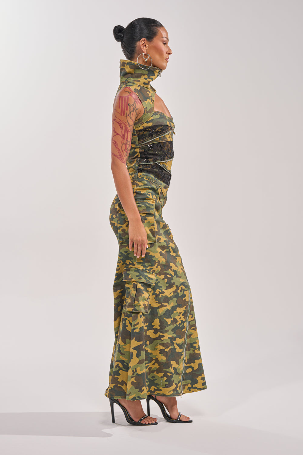 A woman stands in profile wearing the FUSED TOGETHER MOCK NECK MAXI DRESS IN CAMO with black detailing, black high-heeled sandals, large hoop earrings, and her hair styled in a bun. 5