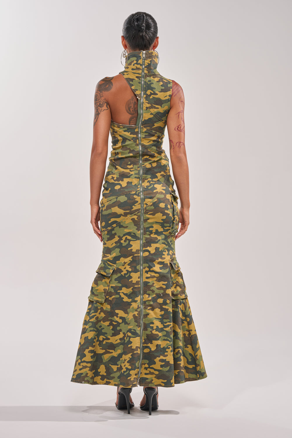 A person faces away in the FUSED TOGETHER MOCK NECK MAXI DRESS IN CAMO, featuring a zippered mock neck, fitted silhouette, utility details, flared hem, visible arm tattoos, and black heels. 6