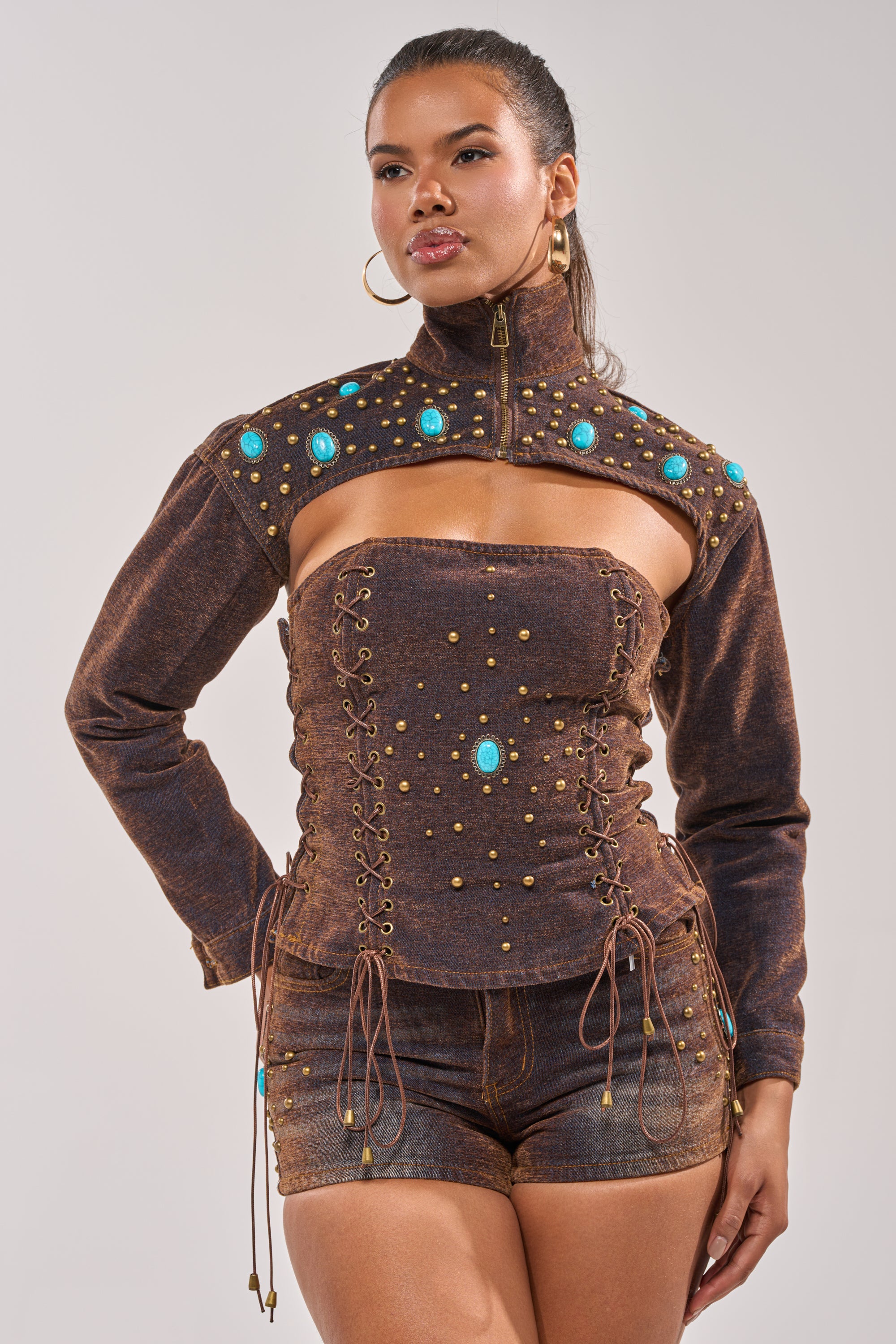 A woman confidently poses against a plain background, wearing the INDIGO STUDDED DENIM BOLERO over a brown off-shoulder corset top with gold studs, turquoise accents, matching high-waisted shorts, and gold hoop earrings. 2